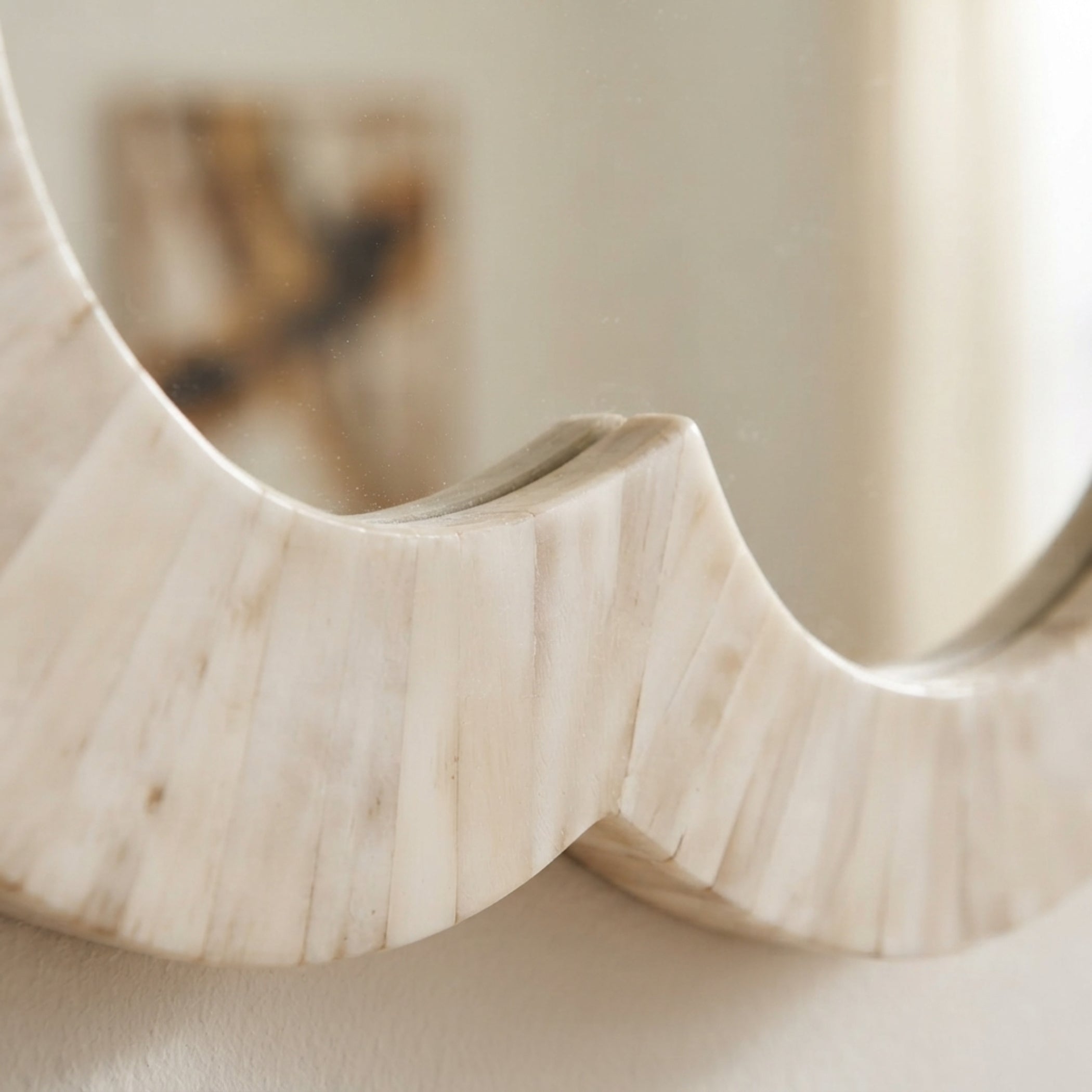 A close-up of a sculptural wavy mirror with a carved bone or ivory frame showing a softly blurred neutral-toned reflection.