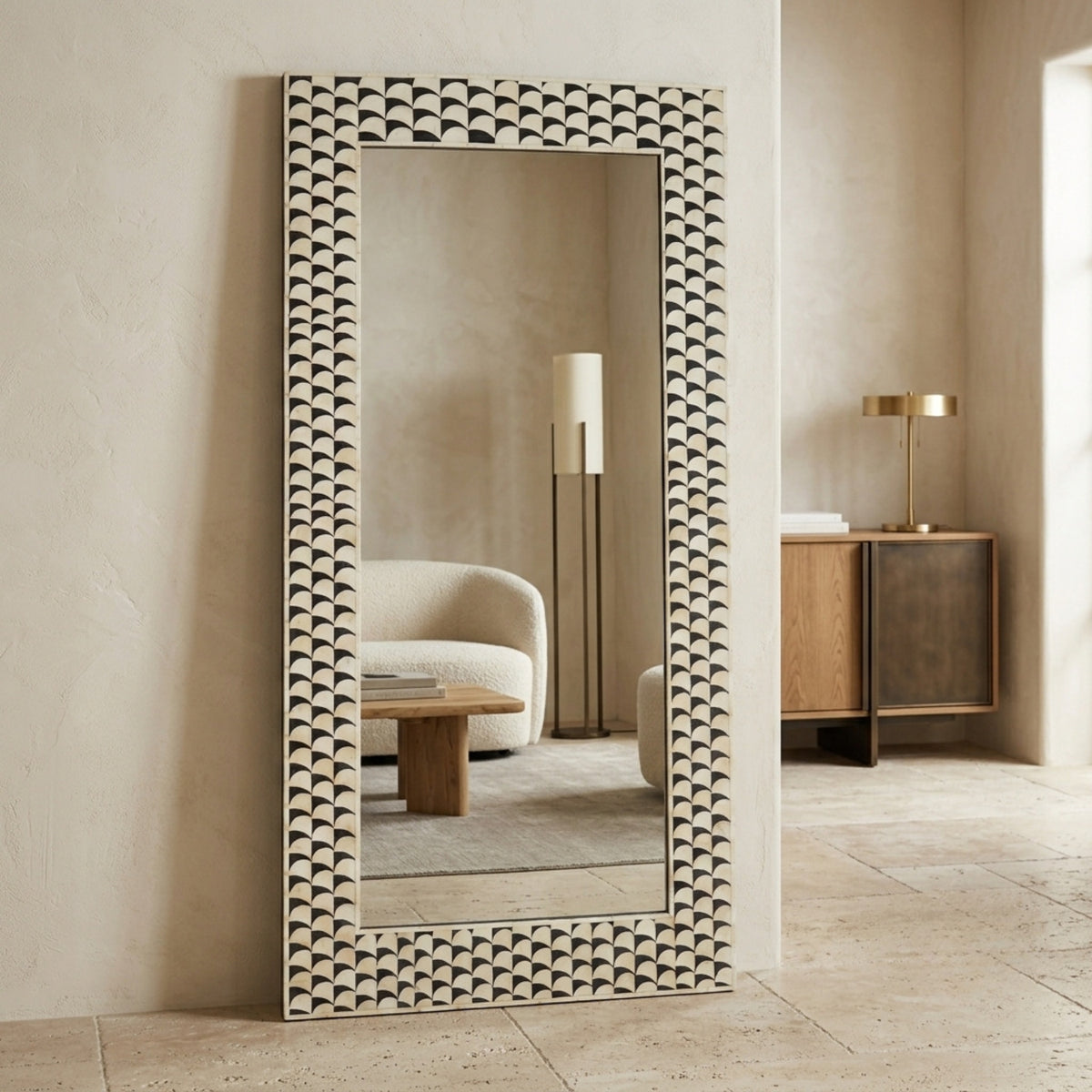 A tall rectangular floor mirror with a decorative scalloped frame, leaning against a beige wall in a styled interior room.