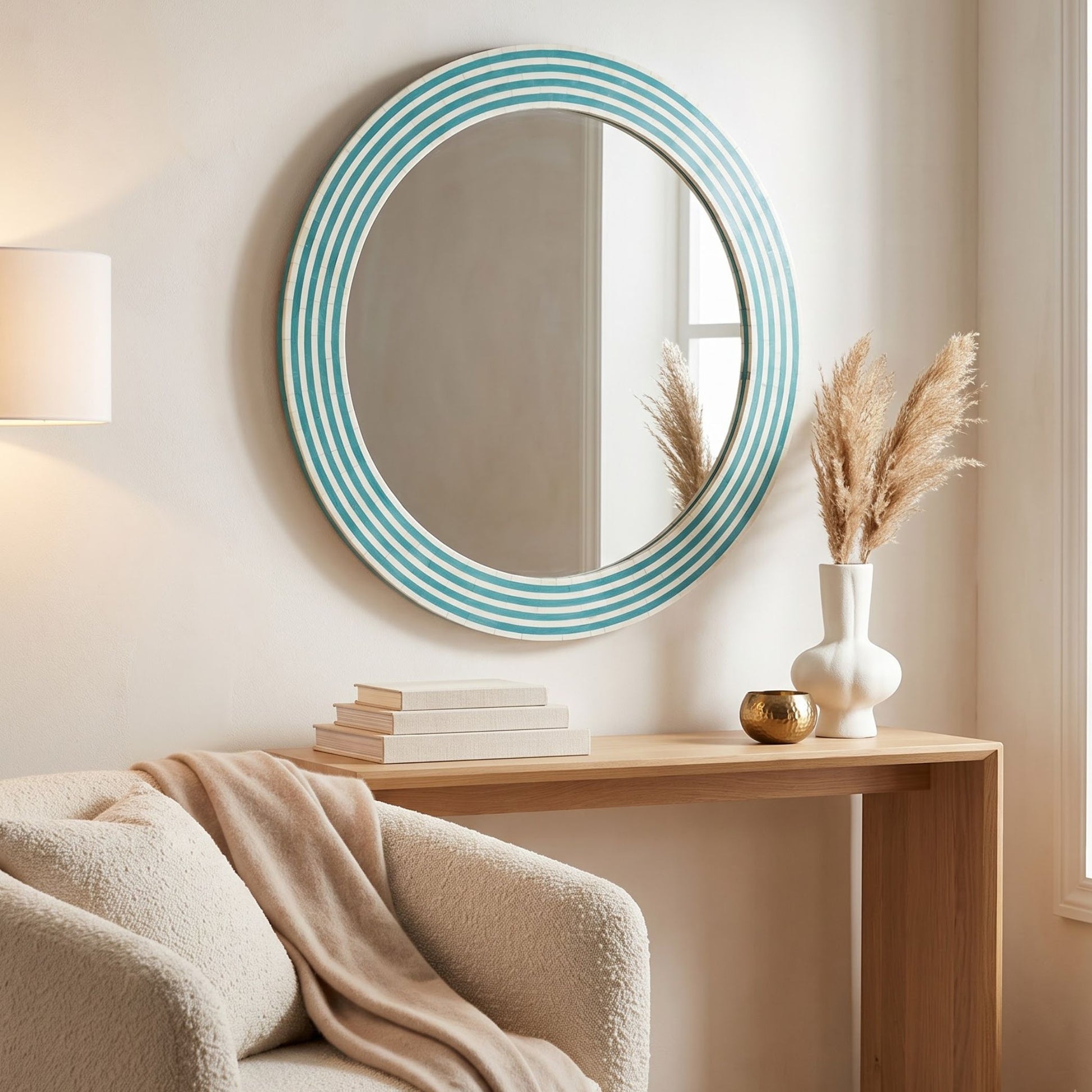 Round wall mirror with a turquoise and white striped frame, styled on a beige wall above a light wood console table with books, a white vase with dried grass, and a cream bouclé chair.