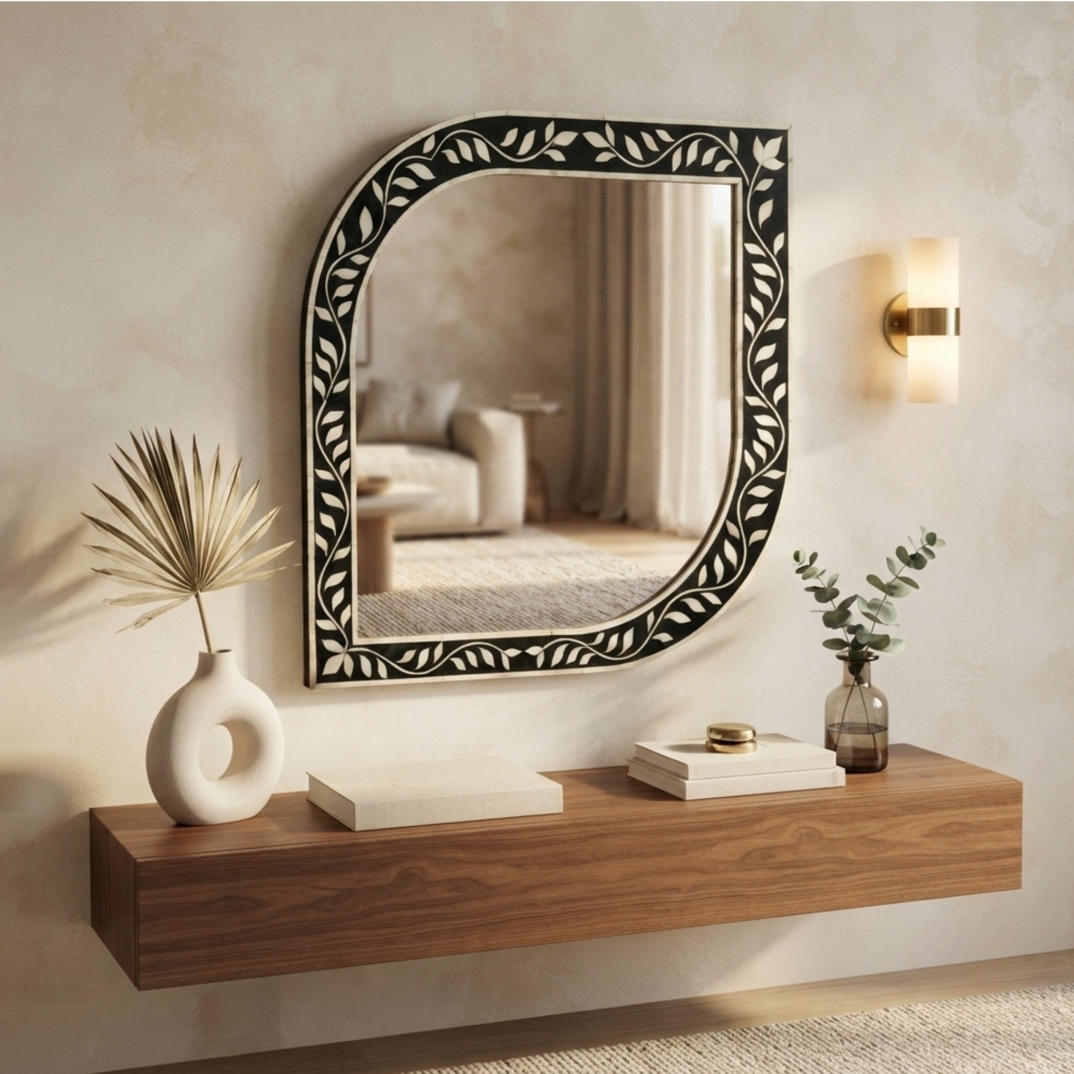 An arched wall mirror with an ornate black and white leaf-patterned frame, mounted on a textured beige wall above a floating wooden console shelf. A warm wall sconce glows to the right, and decorative vases and books sit on the shelf.