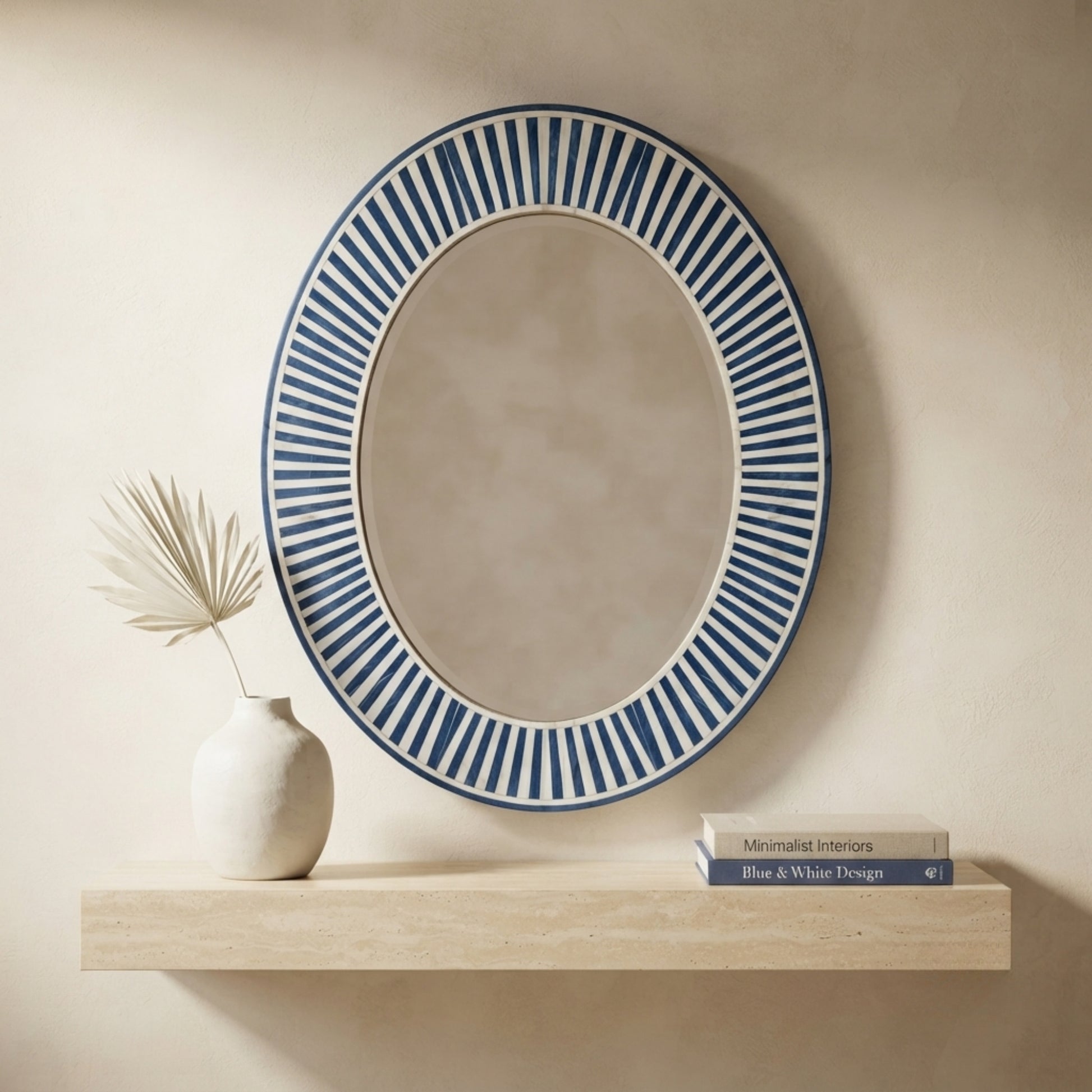 An oval wall mirror with a blue and white striped sunburst border, styled on a light wood floating shelf next to a small white vase with dried palm leaves and a stack of books against a beige wall.