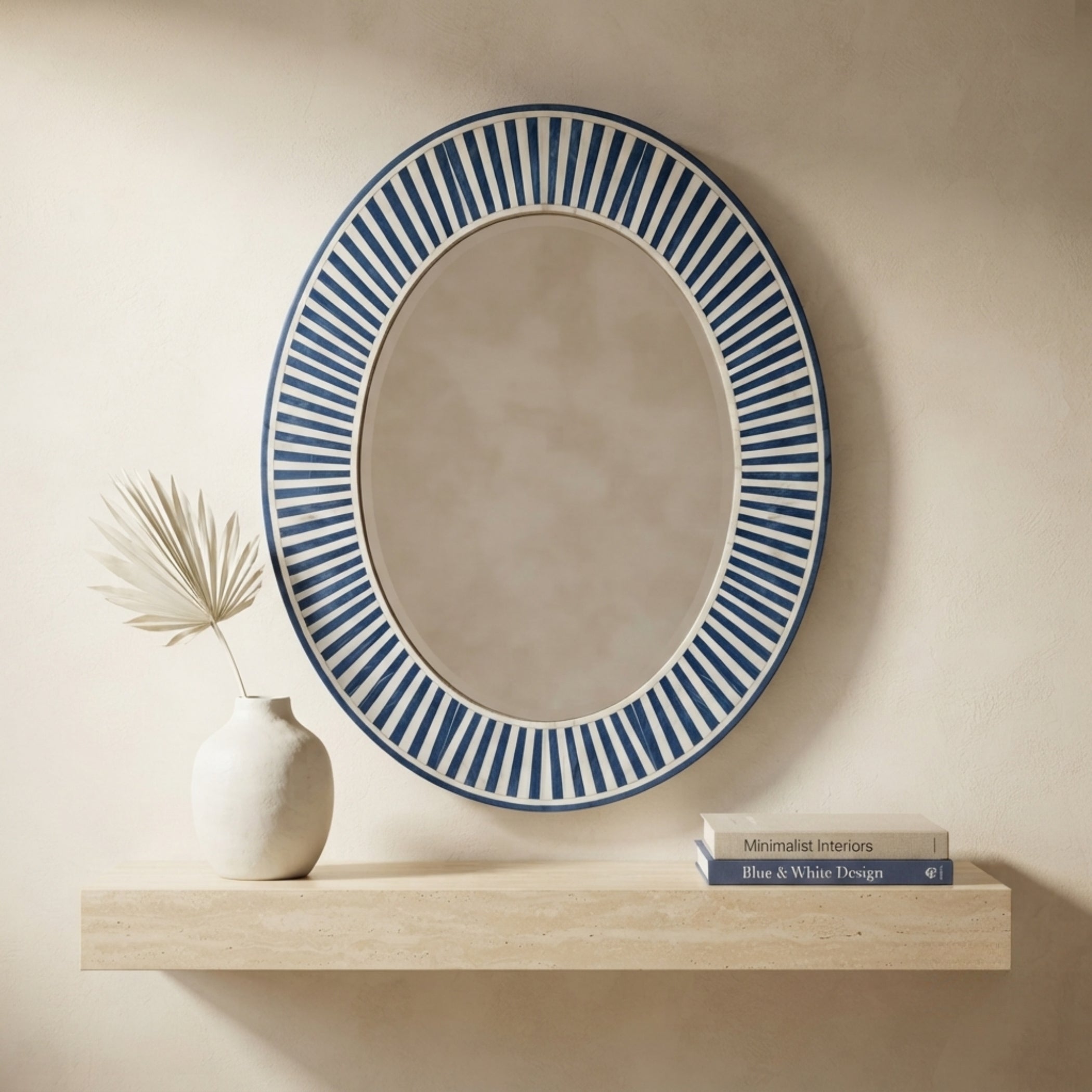 An oval wall mirror with a blue and white striped sunburst border, styled on a light wood floating shelf next to a small white vase with dried palm leaves and a stack of books against a beige wall.