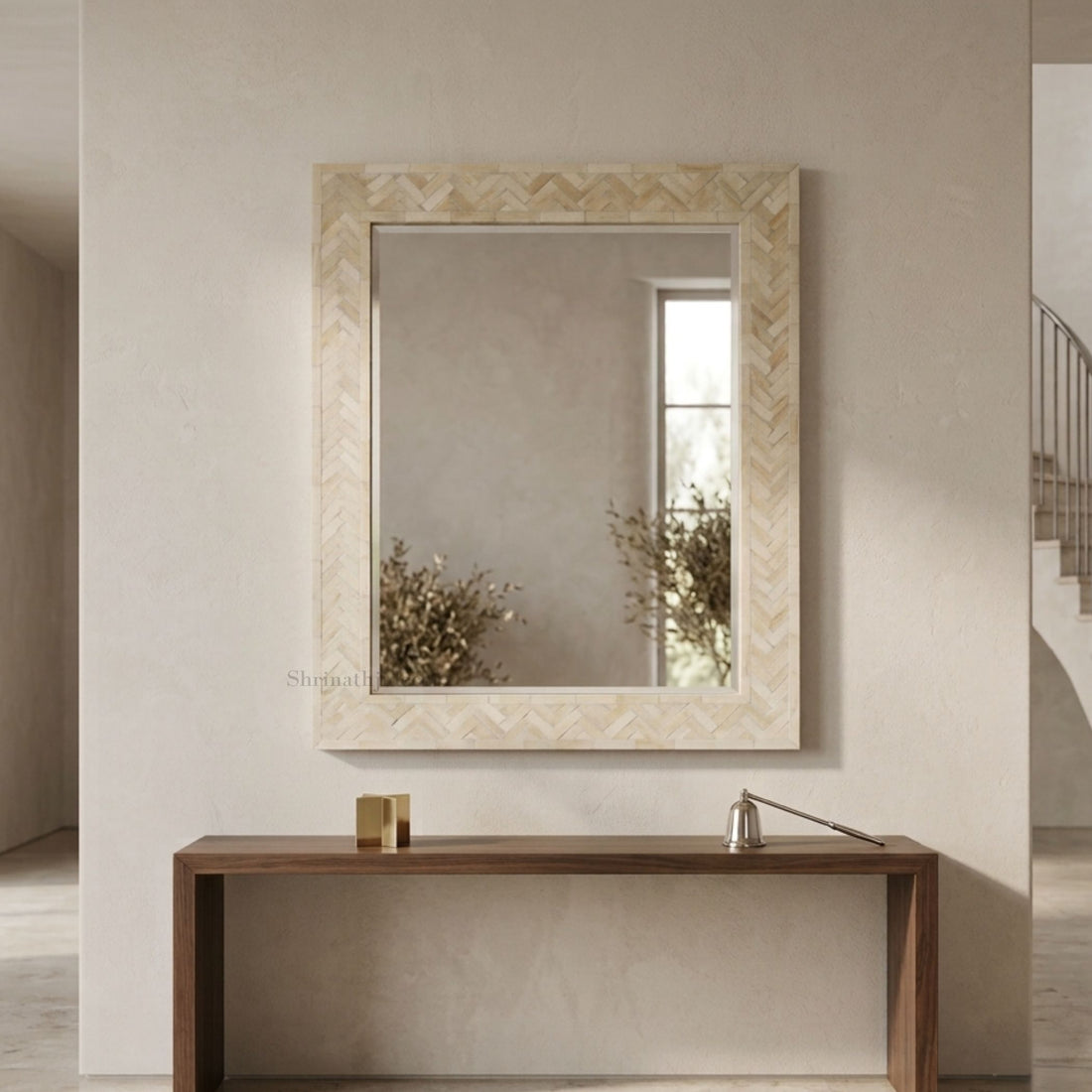 A rectangular wall mirror with a light beige herringbone-patterned frame, mounted on a textured beige wall above a dark wood console table. The mirror reflects a window and dried plants, and a small brass lamp sits on the table.