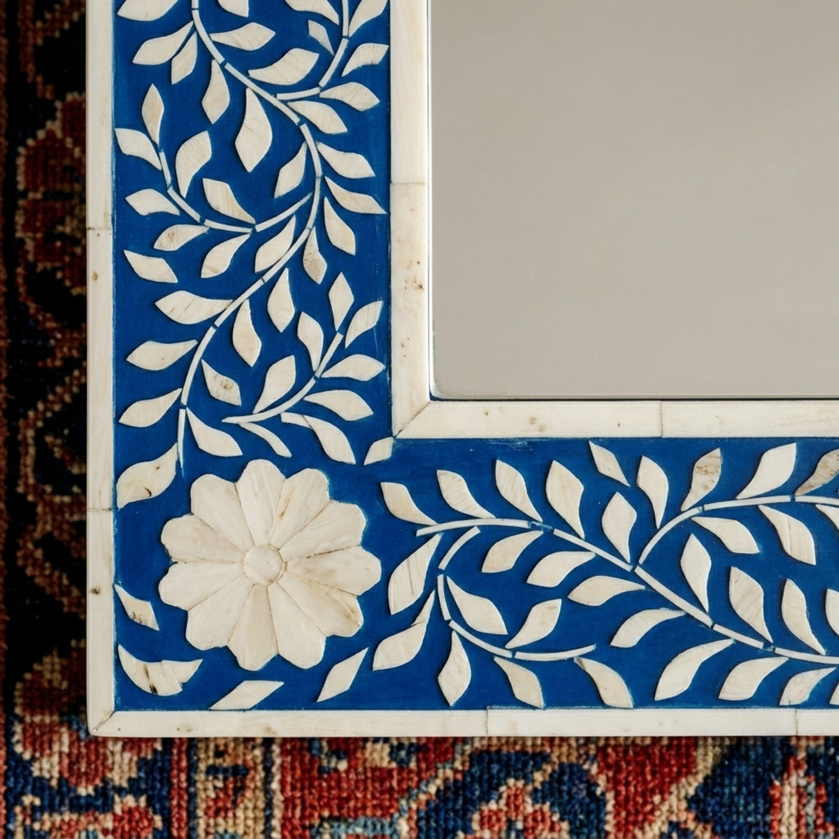 A close-up of a decorative mirror frame featuring intricate white bone or mother-of-pearl floral and leaf inlay on a vibrant blue background, with a large sculpted flower in the lower left corner.