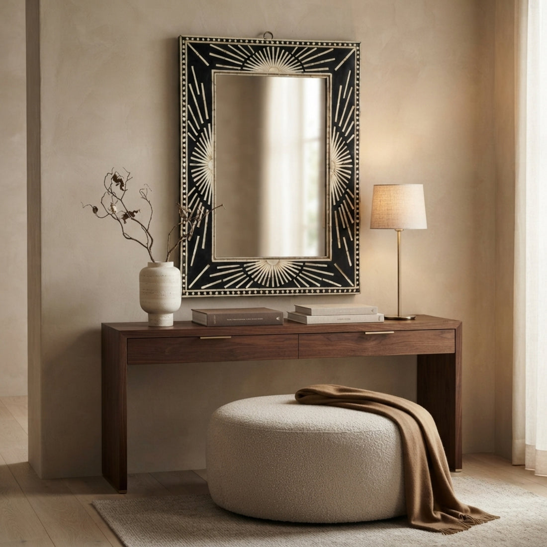 A large rectangular wall mirror with an ornate black and gold sunburst frame hangs above a dark wood console table in a styled room. A white ceramic vase with dried branches, stacked books, a table lamp, and a round textured ottoman with a draped brown throw are arranged below.