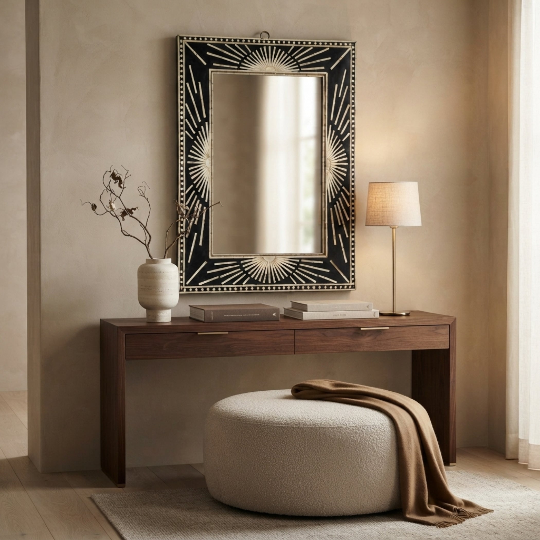 A large rectangular wall mirror with an ornate black and gold sunburst frame hangs above a dark wood console table in a styled room. A white ceramic vase with dried branches, stacked books, a table lamp, and a round textured ottoman with a draped brown throw are arranged below.