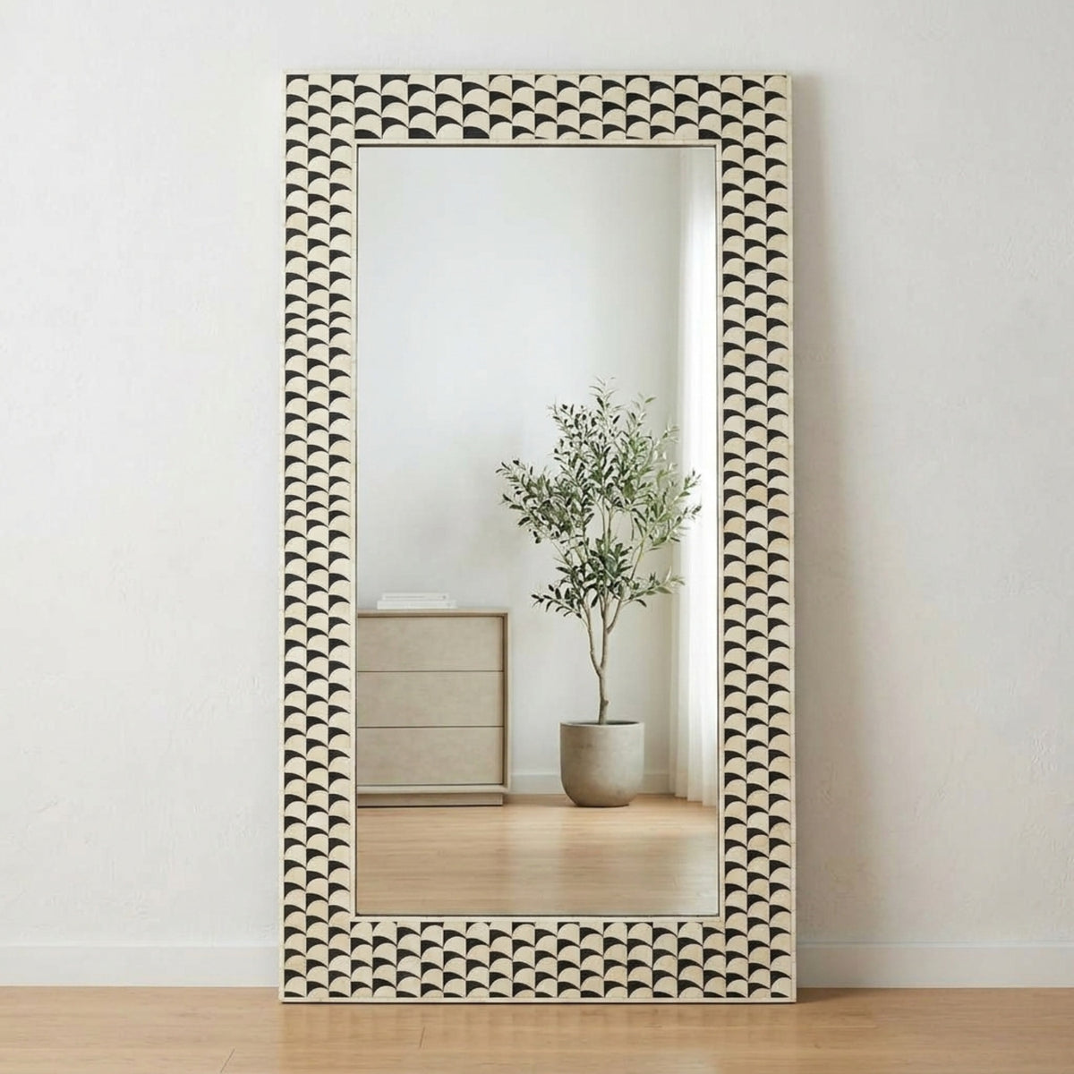 A tall rectangular floor mirror with an intricate hand-carved frame featuring a repeating fan or scallop pattern in cream and dark brown, leaning against a white wall on a light wood floor. The mirror reflects a potted olive tree and a wooden cabinet.