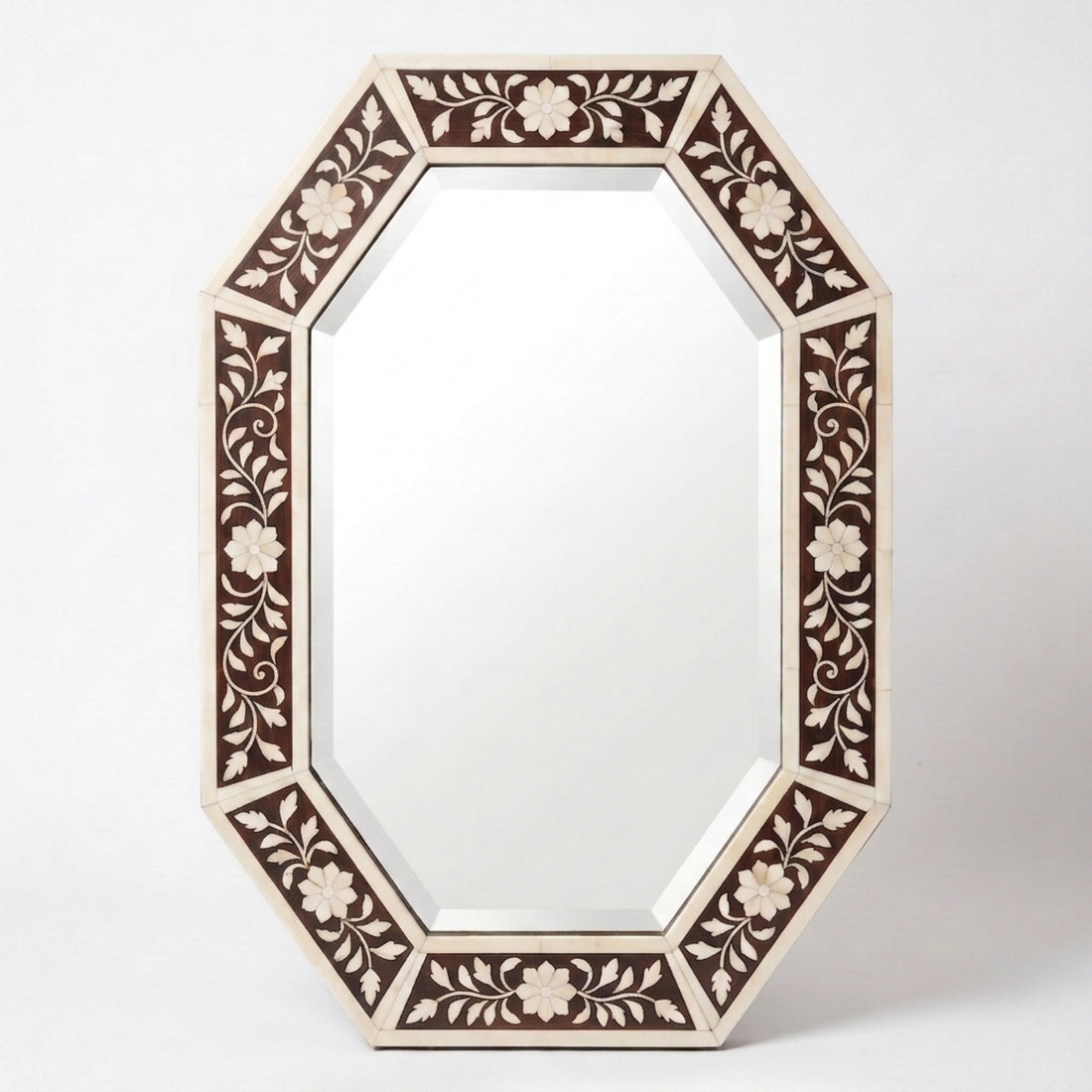 An octagonal wall mirror with an intricate carved floral frame in cream and dark brown, mounted on a textured beige wall above a wooden console table with books and a vase.