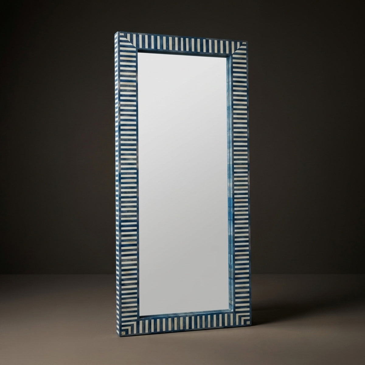 A tall rectangular wall mirror with a decorative striped frame in blue and white, standing against a dark gray studio background.