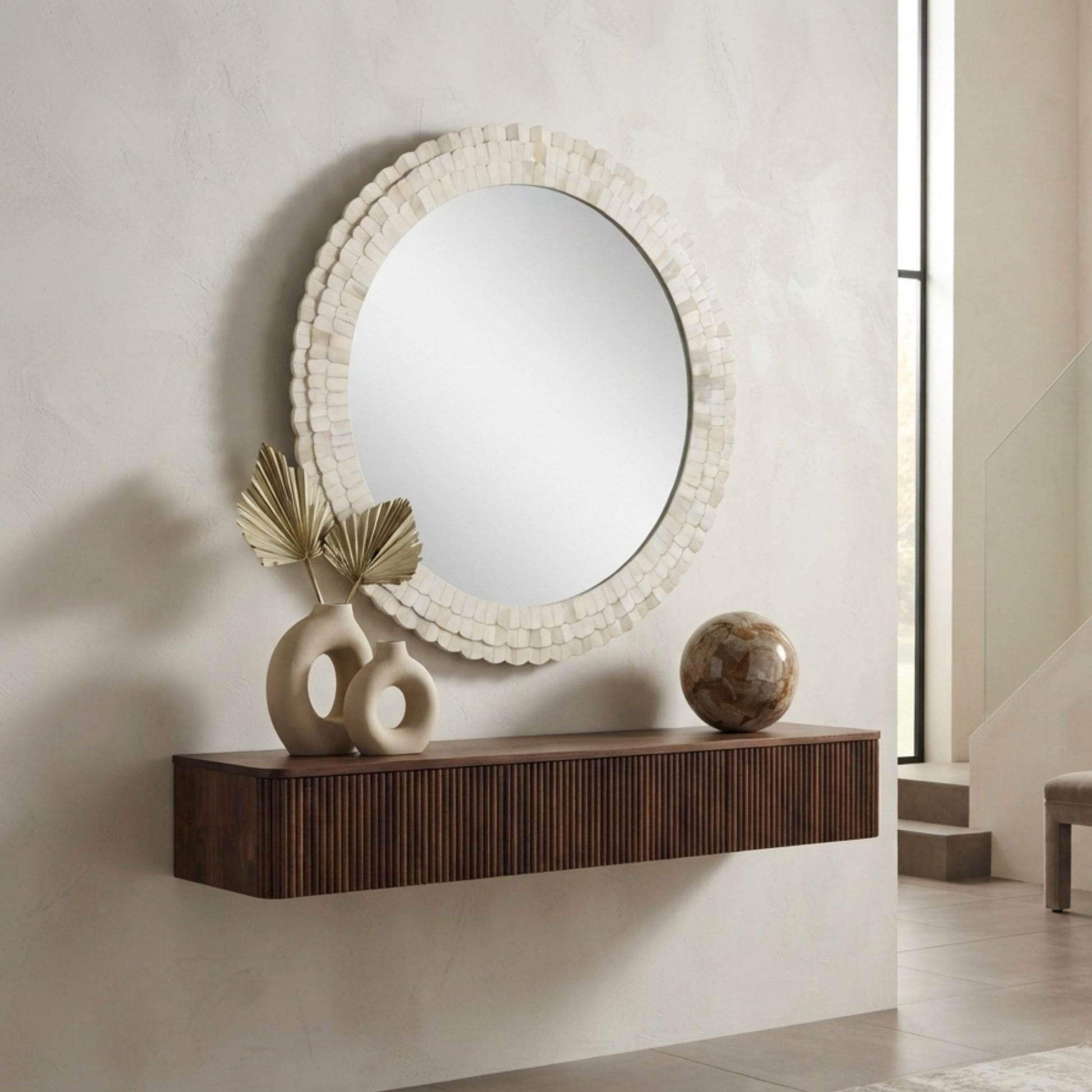 An oval wall mirror with a textured, layered off-white frame hangs above a floating dark brown wooden console shelf in a modern interior setting. Decorative ceramic vases and a stone orb sit on the shelf against a textured beige wall.