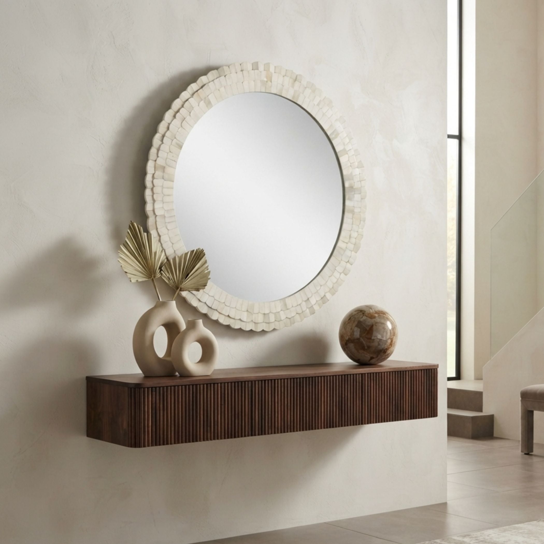 An oval wall mirror with a textured, layered off-white frame hangs above a floating dark brown wooden console shelf in a modern interior setting. Decorative ceramic vases and a stone orb sit on the shelf against a textured beige wall.