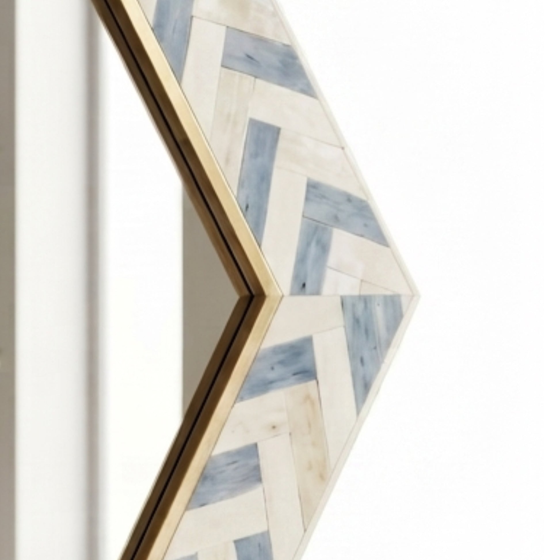 A close-up of a geometric arrow-shaped wall mirror with a gold frame and a herringbone wood inlay pattern in off-white and light blue, shown against a white background.