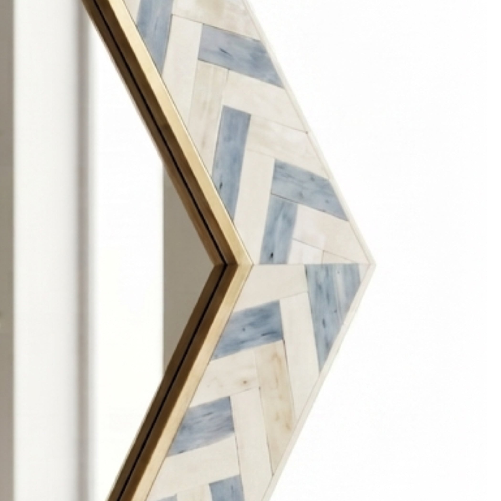 A close-up of a geometric arrow-shaped wall mirror with a gold frame and a herringbone wood inlay pattern in off-white and light blue, shown against a white background.