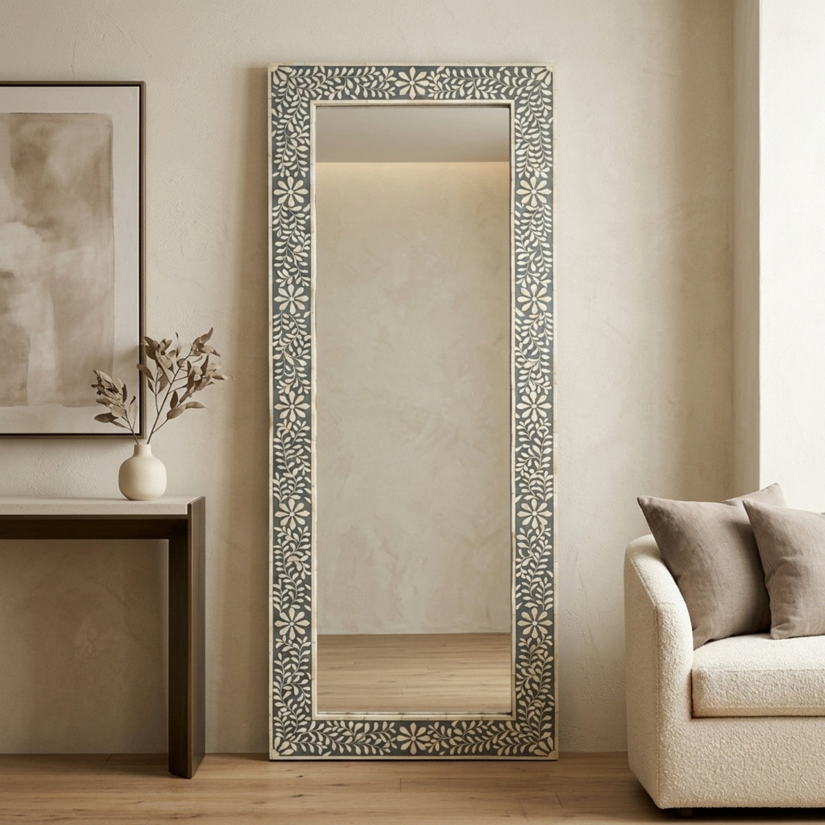 A tall rectangular floor mirror with an ornate carved or inlaid floral border in gray and white, styled in a modern living room next to a sofa and console table.
