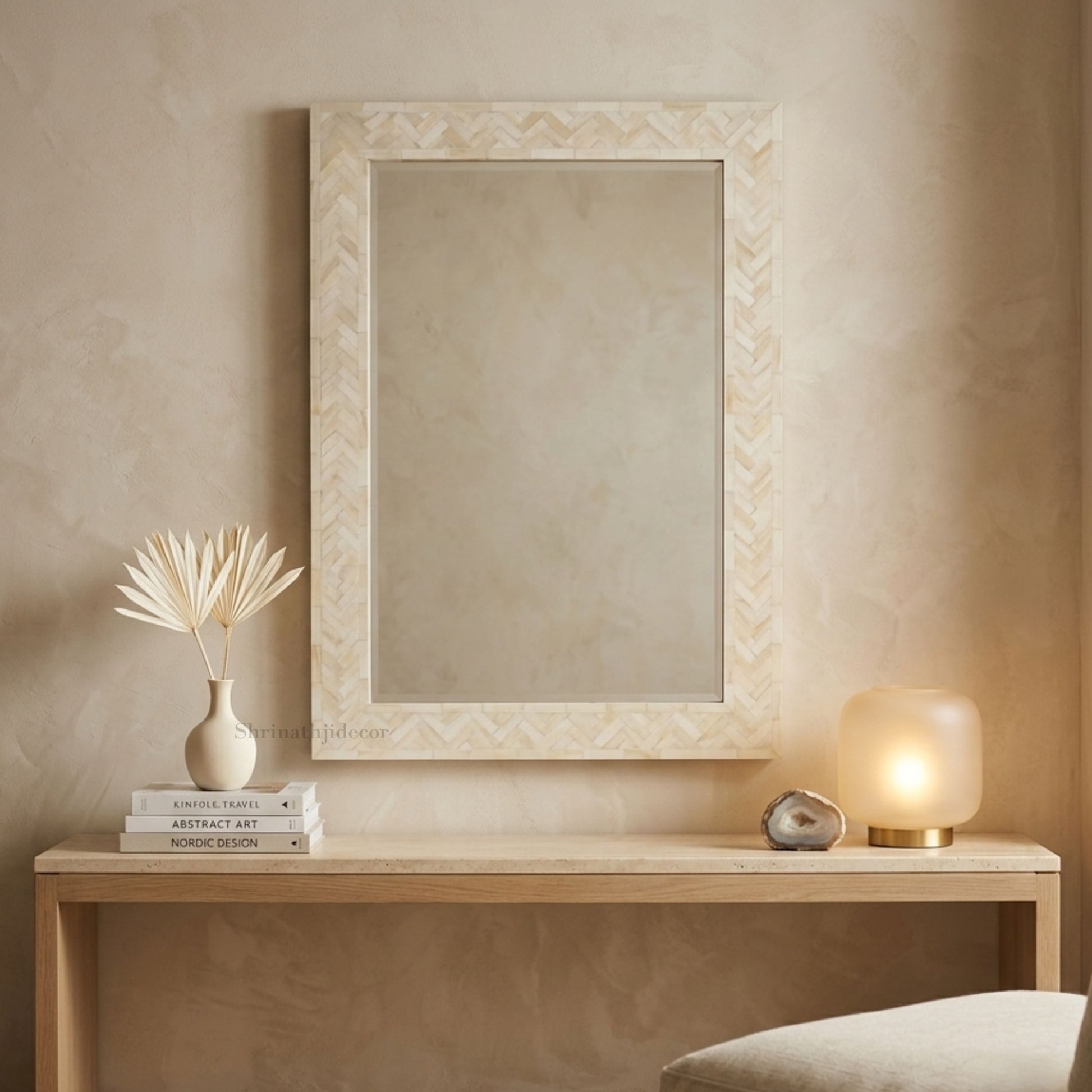 A rectangular wall mirror with a cream-colored chevron-patterned frame hangs on a textured beige wall above a light wood console table styled with a vase, books, and a glowing lamp.