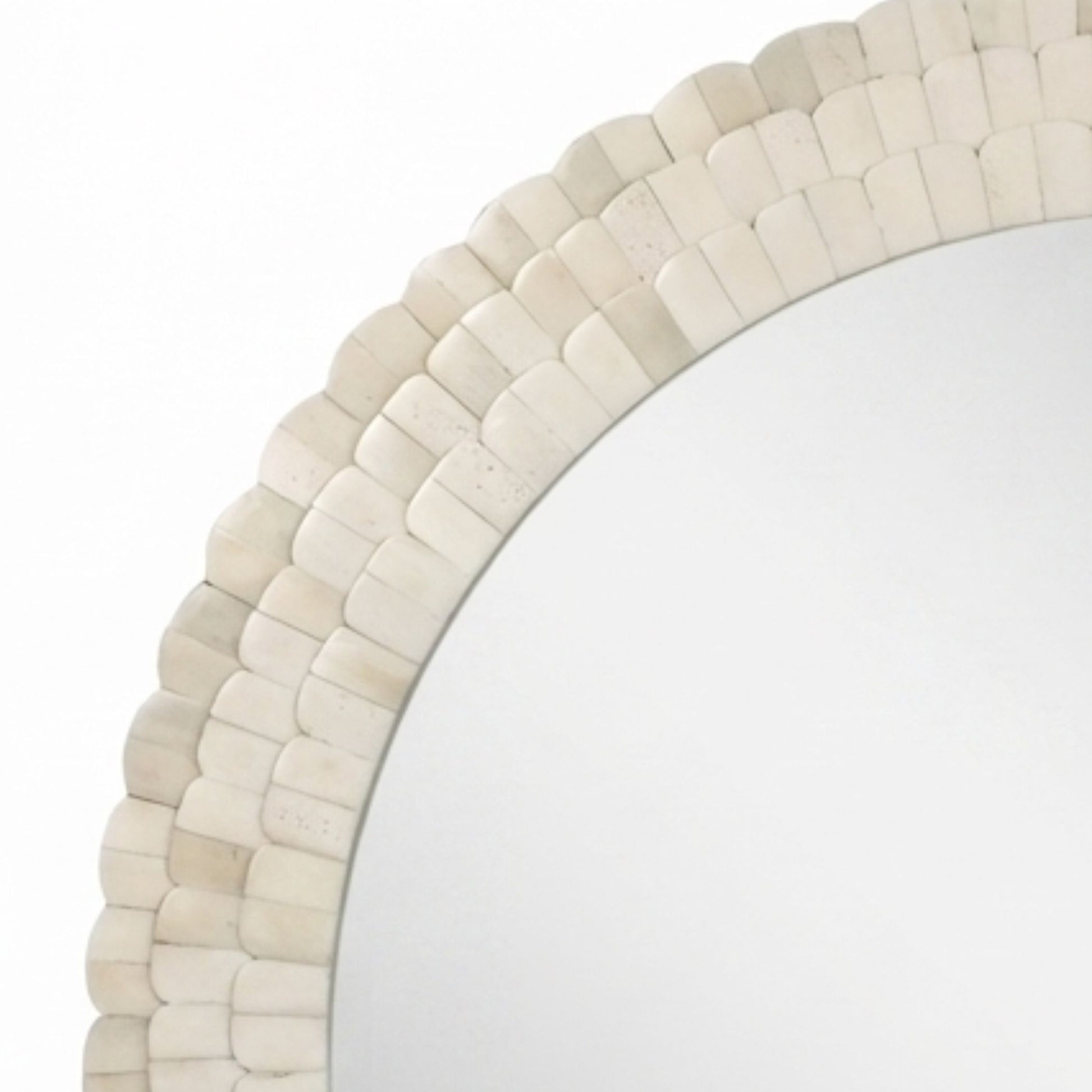 A close-up of a round mirror with a decorative scalloped frame made of small overlapping shell-like pieces in an off-white or cream tone, shown against a white background.