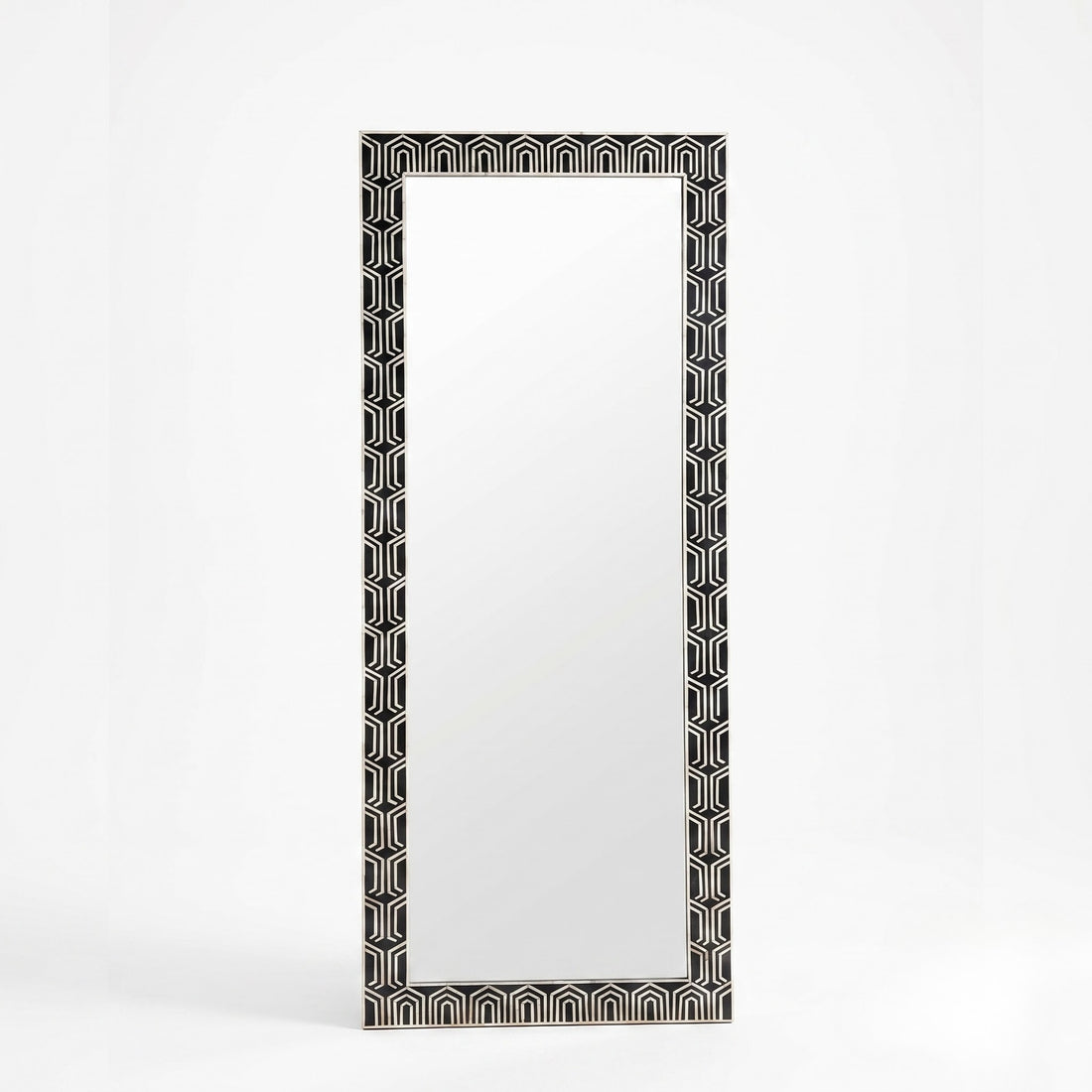A tall rectangular floor mirror with a black and white geometric patterned frame, styled in a warm, modern room next to a console table and a floor lamp.