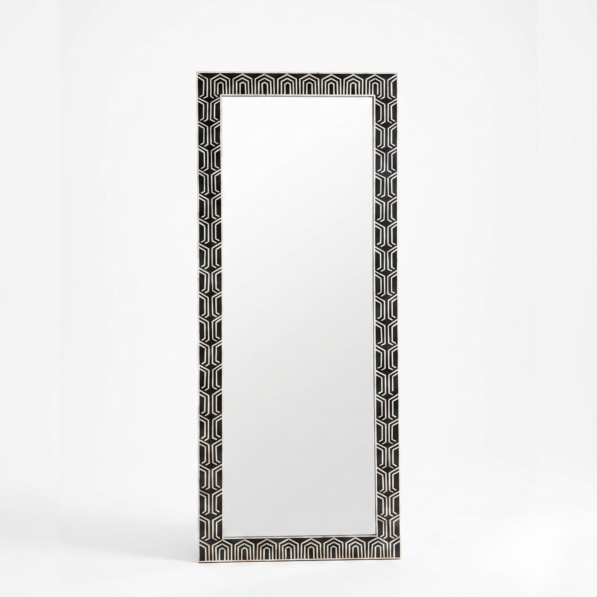 A tall rectangular wall mirror with a black and white geometric patterned frame, standing against a clean white background.