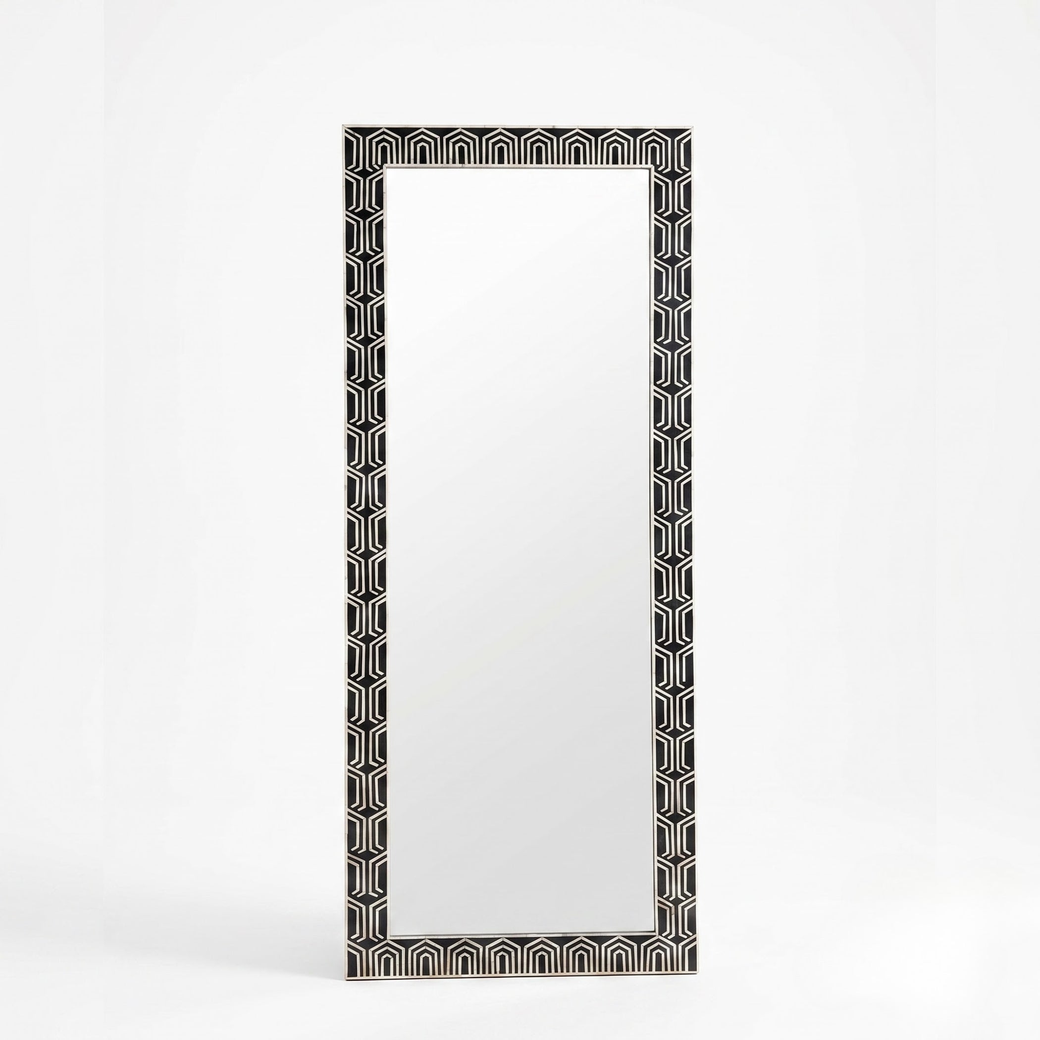 A tall rectangular wall mirror with a black and white geometric patterned frame, standing against a clean white background.