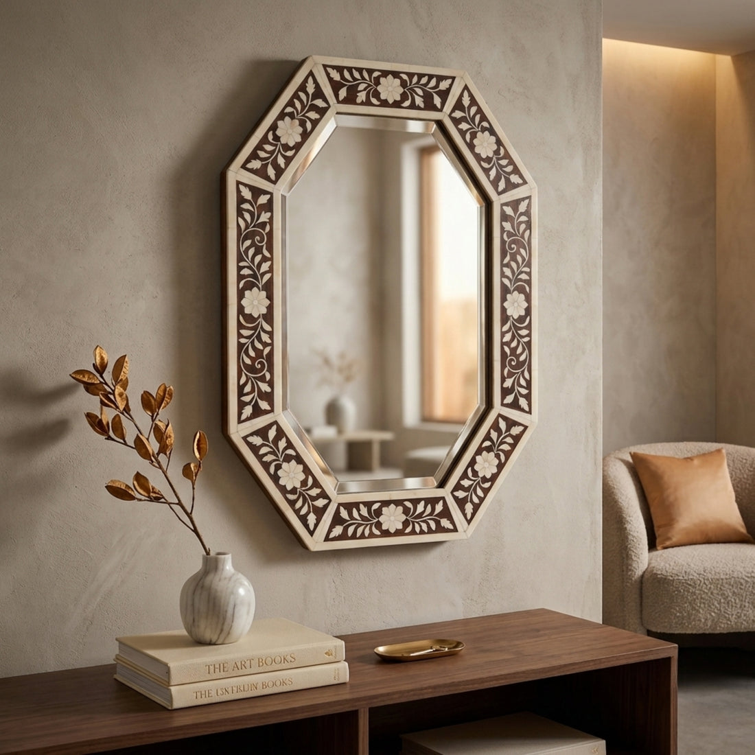 An octagonal wall mirror with an intricate carved floral frame in cream and dark brown, mounted on a textured beige wall above a wooden console table with books and a vase.