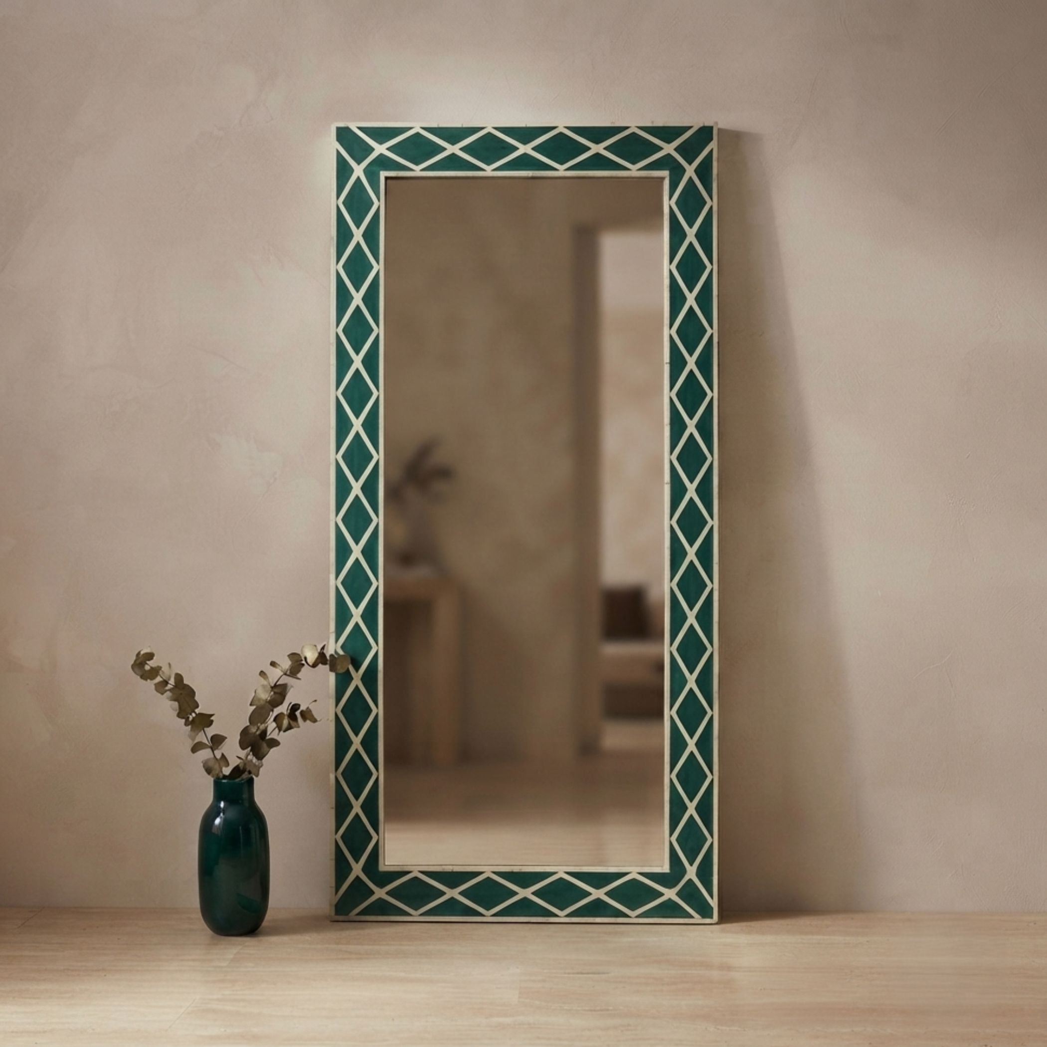 A tall rectangular wall mirror with a decorative geometric border in dark teal and cream, styled on a light wood floor against a textured beige wall next to a dark teal vase with dried branches.