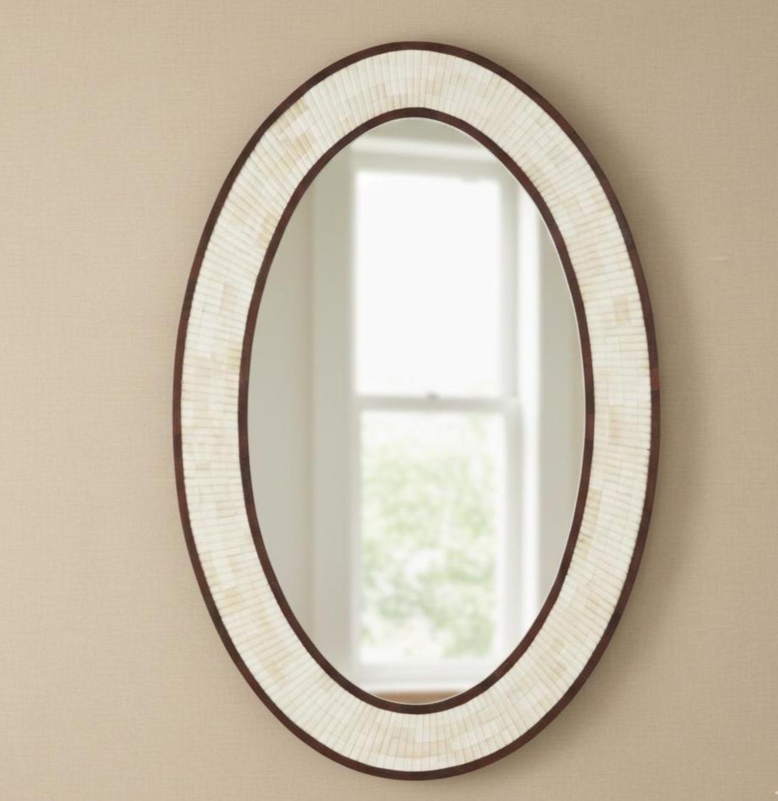 An oval wall mirror with a dark brown frame and a textured cream-colored shell or tile border, mounted on a beige wall with a window reflection visible in the glass.