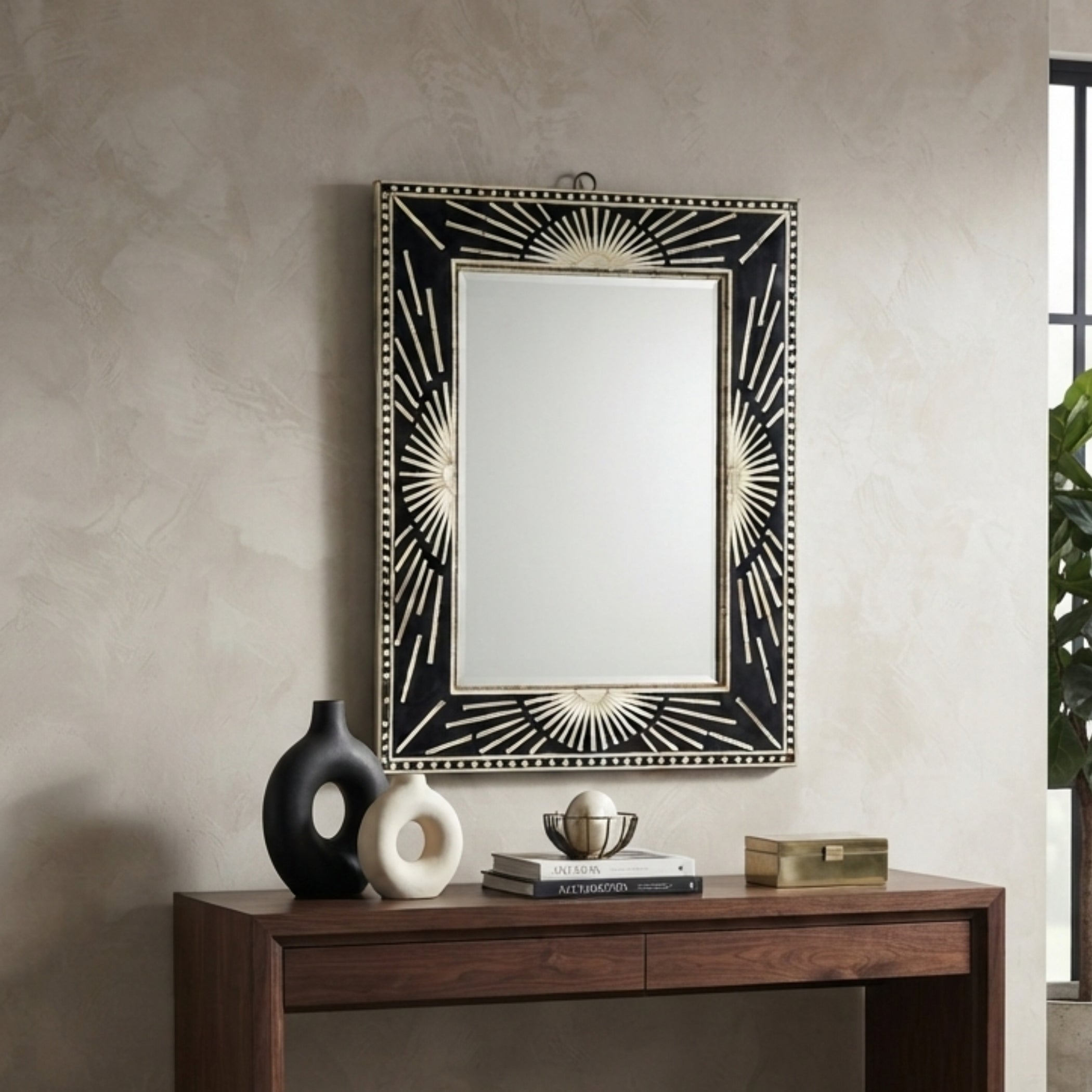A rectangular wall mirror with an ornate black and gold sunburst frame, mounted on a textured beige wall above a dark wood console table with decorative objects.