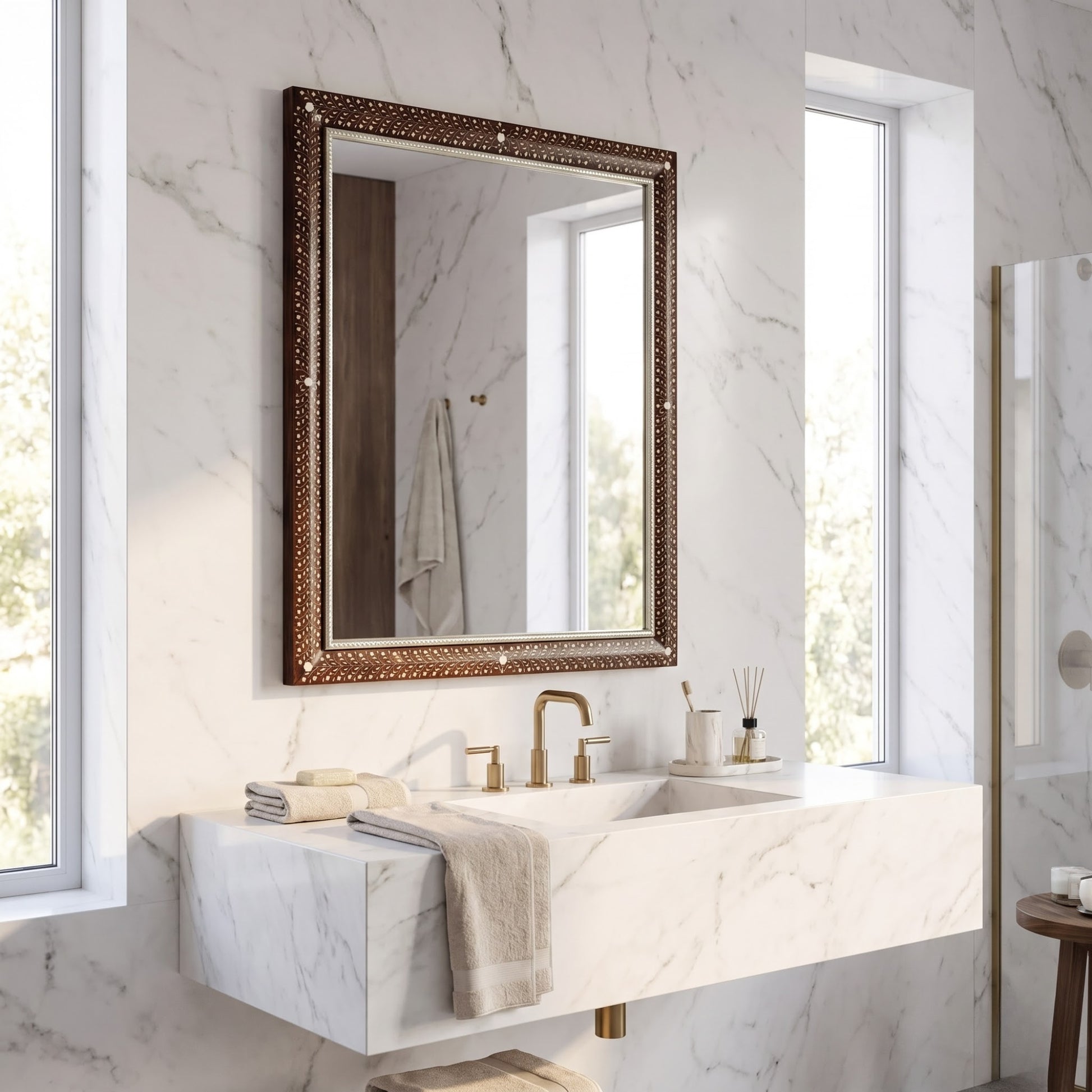 An ornate rectangular bathroom mirror with a textured bronze frame hangs above a white marble sink and countertop in a bright, modern bathroom with marble walls and large windows.