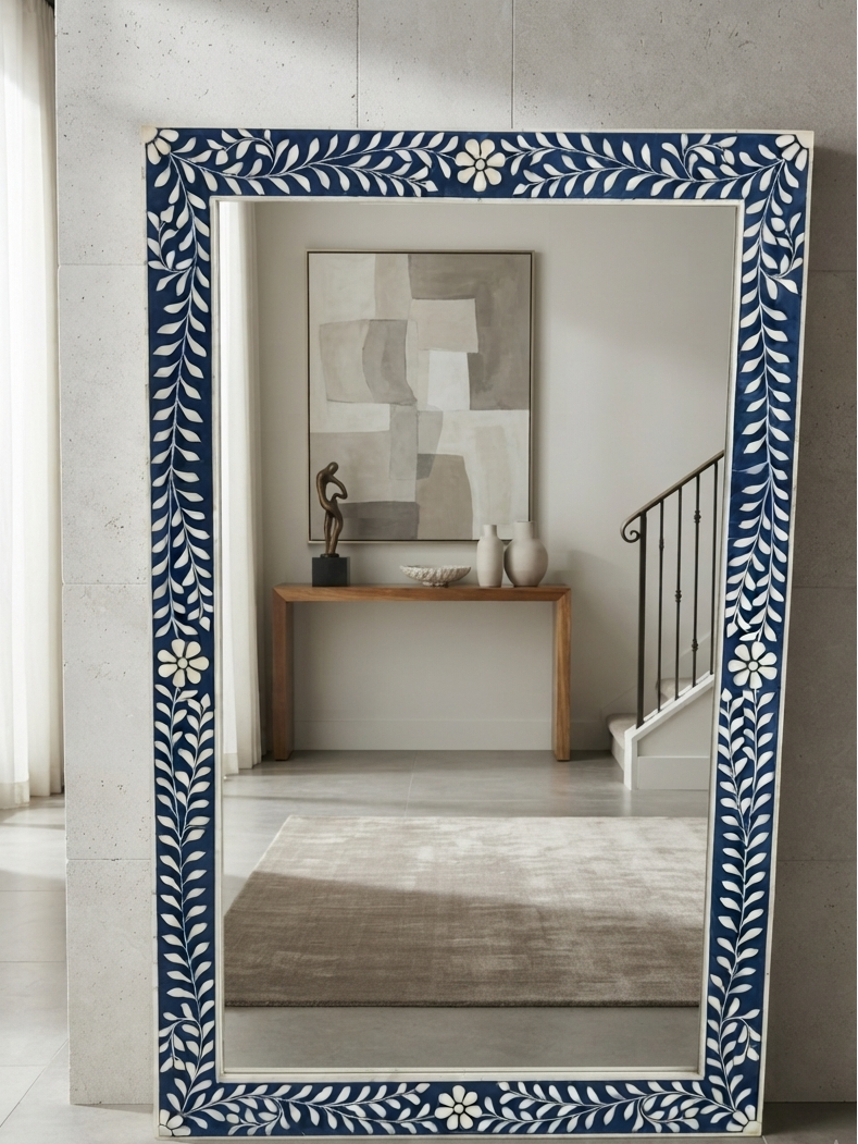 A tall rectangular floor mirror with a decorative navy blue and white floral border, leaning against a textured gray concrete wall in a modern interior setting.