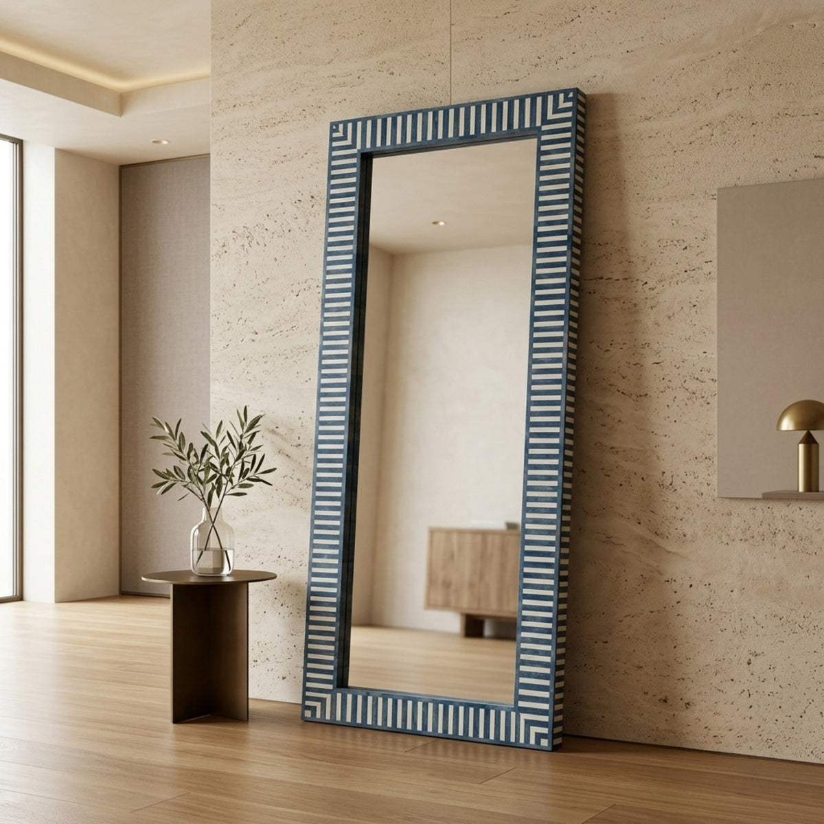 A tall rectangular floor mirror with a blue and white striped frame, leaning against a textured beige stone wall in a modern interior setting.