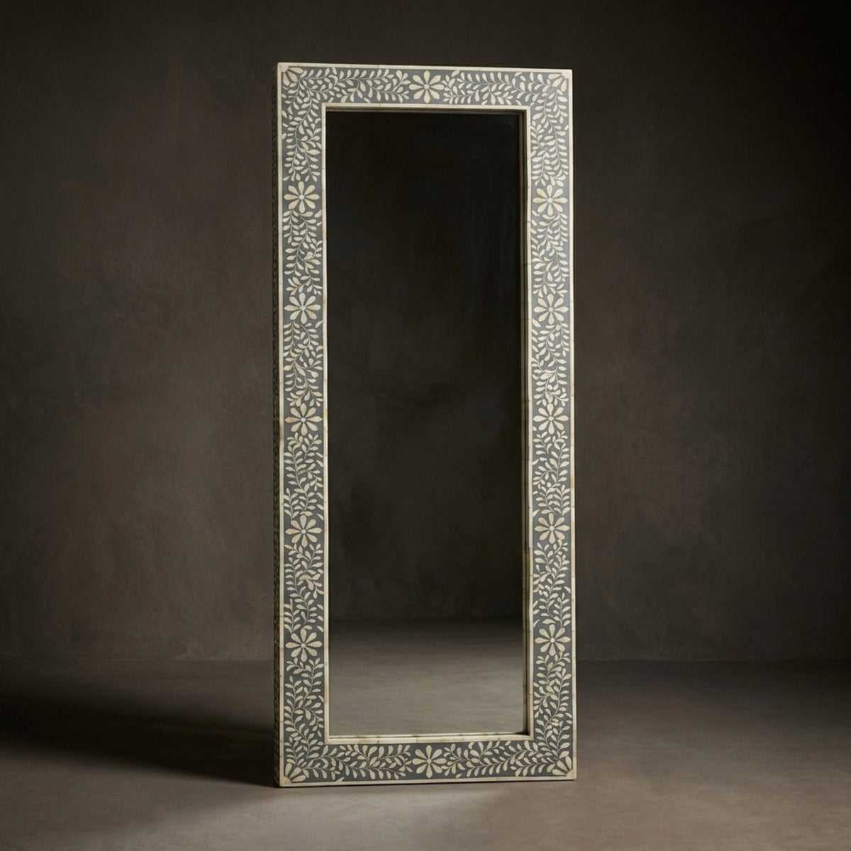 A tall rectangular floor mirror with an intricately carved off-white floral frame, standing against a dark textured studio background.