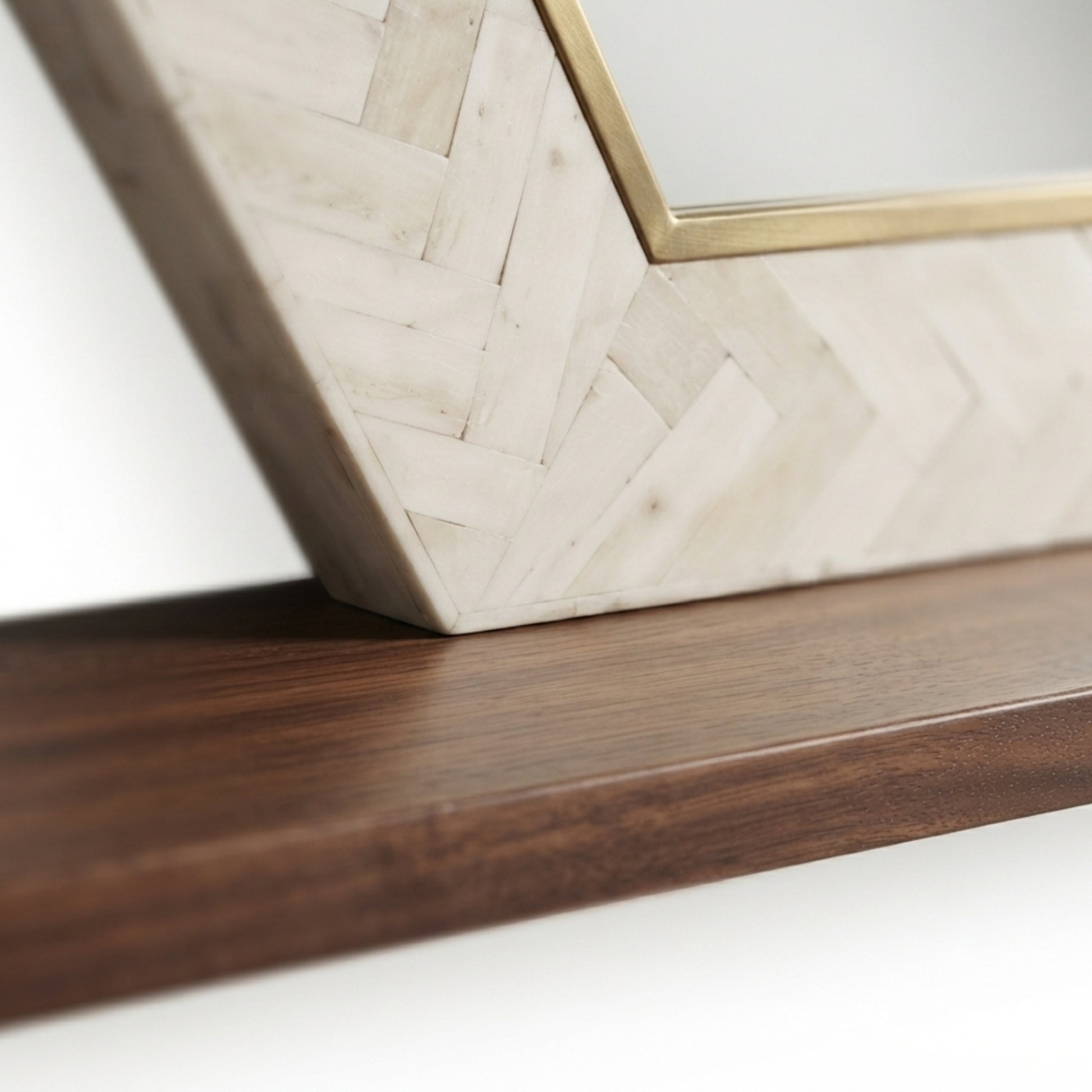 Close-up of a decorative wall mirror with a geometric frame made of light beige herringbone wood, a thin gold metal border, and a dark brown wooden shelf below.