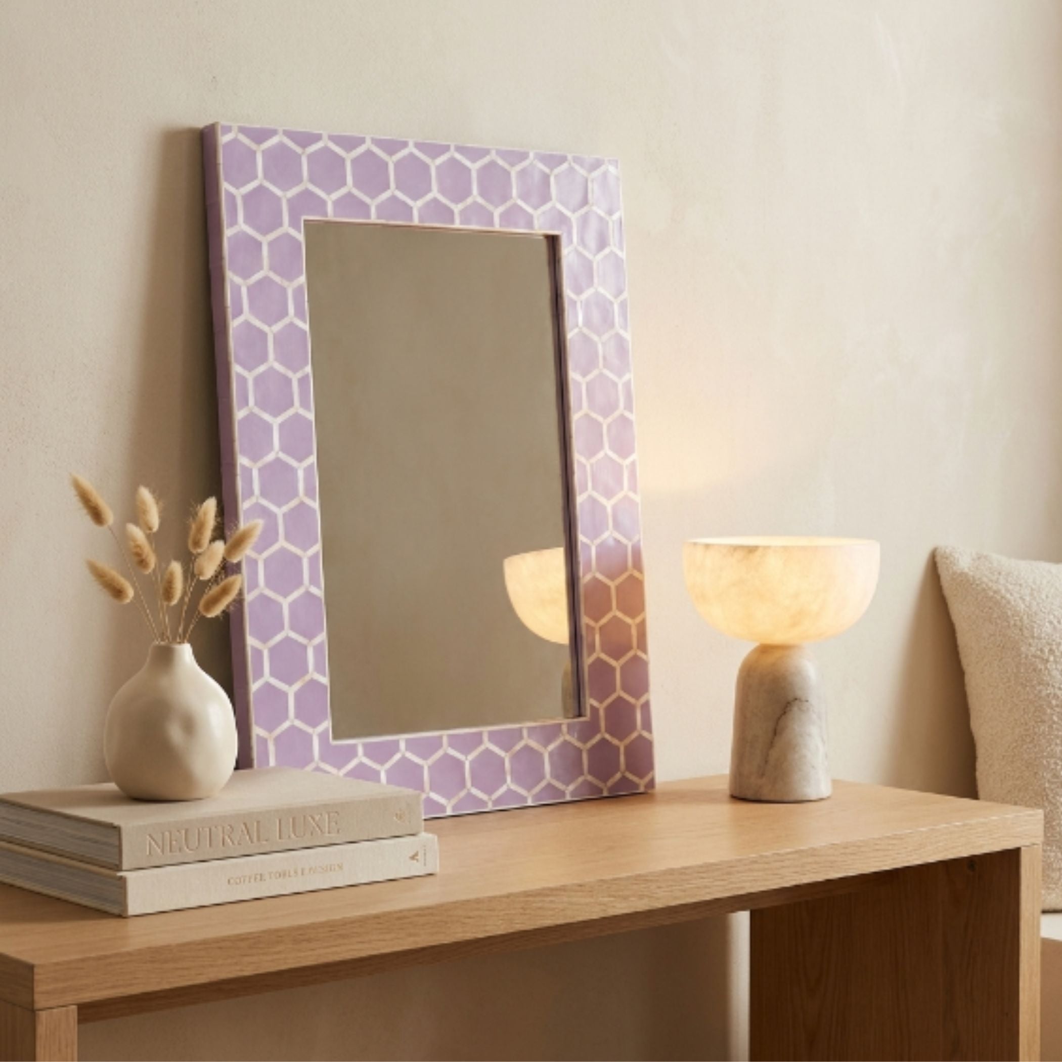 A rectangular wall mirror with a lavender and white geometric honeycomb patterned frame, styled on a light wood console table next to a marble lamp and books.