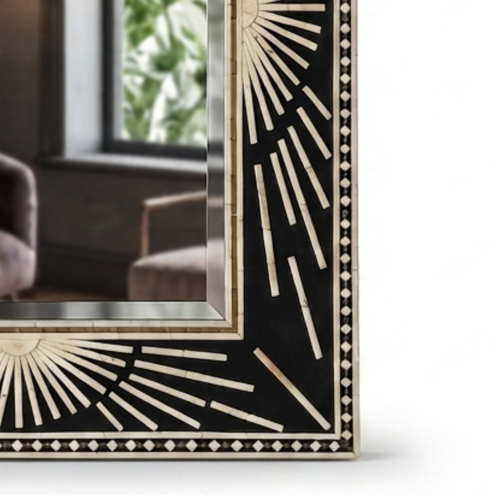 A close-up of a decorative rectangular mirror frame with a black background and beige inlay detailing in a sunburst and geometric pattern, with a blurred interior room reflection visible in the mirror.