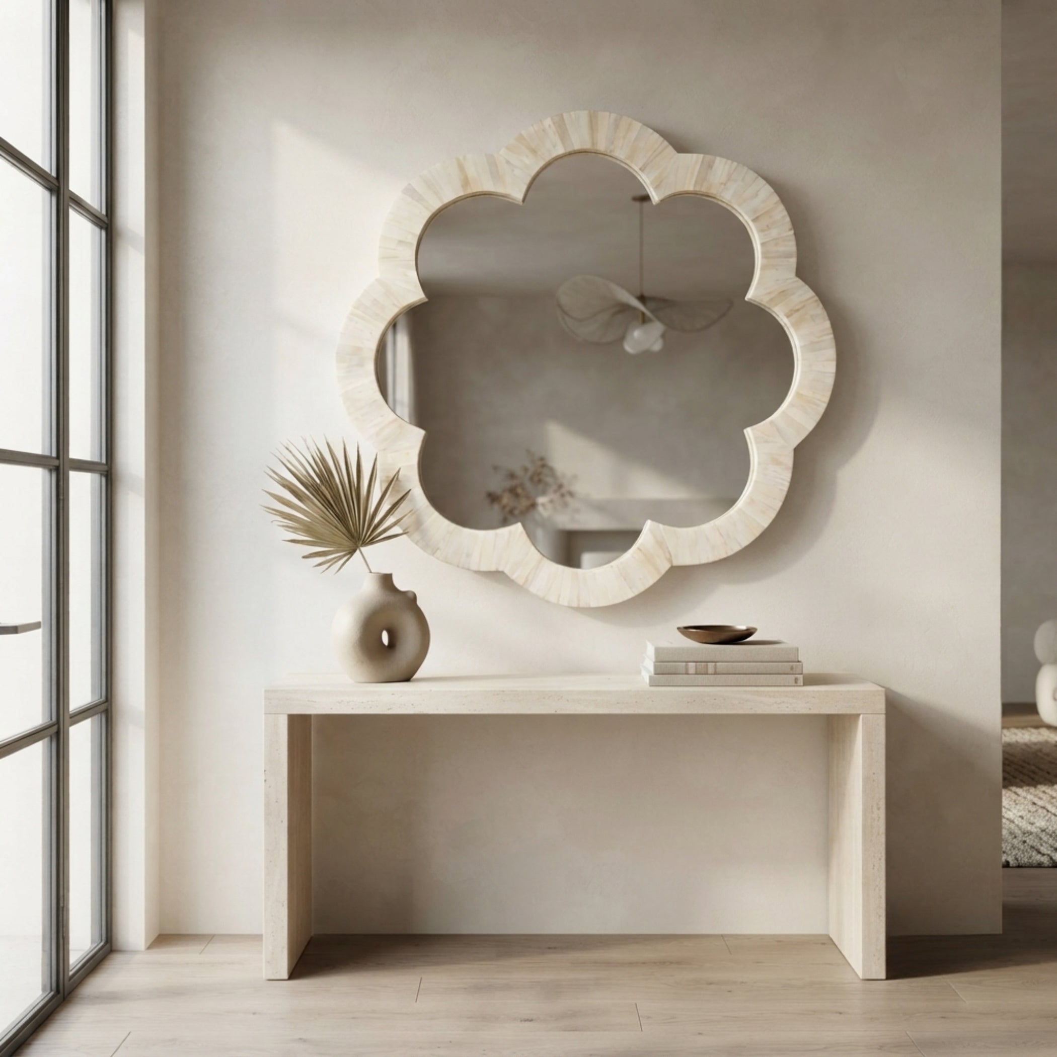 A scalloped cloud-shaped wall mirror with a light natural stone frame hangs above a minimalist console table in a bright, modern room with a large window.