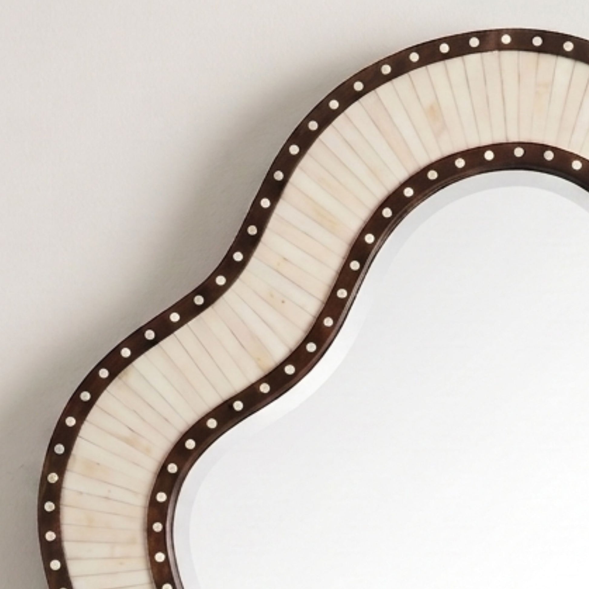 An irregularly shaped wall mirror with a wavy border featuring a dark brown frame, light beige inlay panels, and small white dot accents, shown against a plain light background.