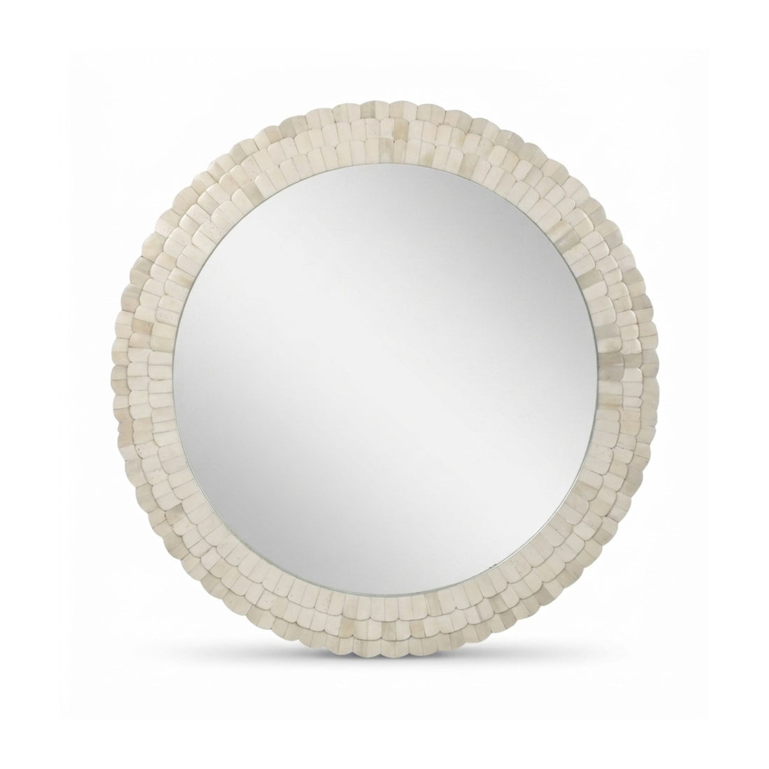 Round wall mirror with a textured cream shell or tile border, mounted above a floating wooden console shelf in a warm neutral room. A beige vase with dried white flowers and stacked books sit on the shelf.