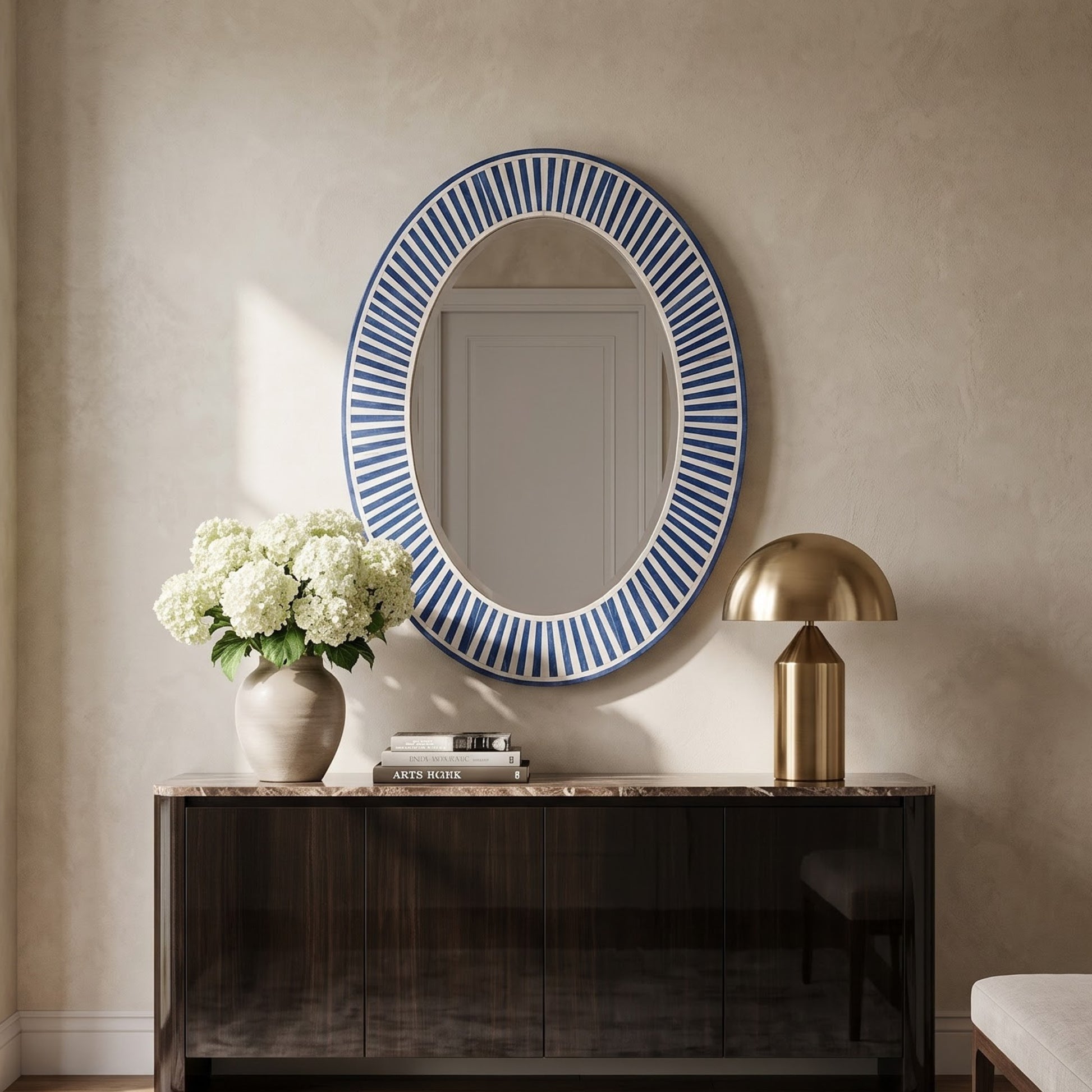 An oval wall mirror with a blue and white striped border, styled in a home interior above a dark wood console table with a vase of white flowers and a gold table lamp.