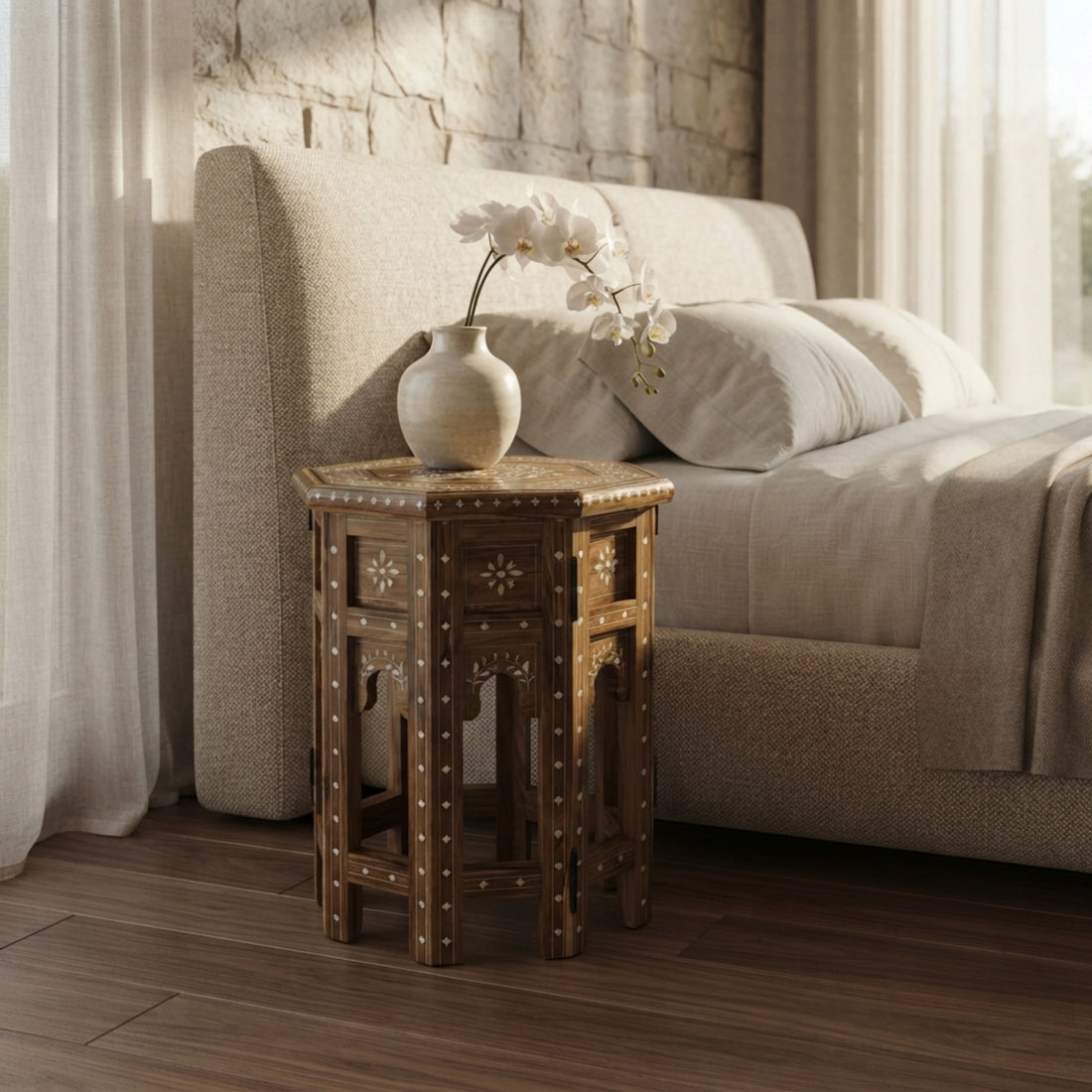 A small octagonal wooden side table with carved details stands beside a bed in a softly lit bedroom. A cream ceramic vase with white orchids rests on top.