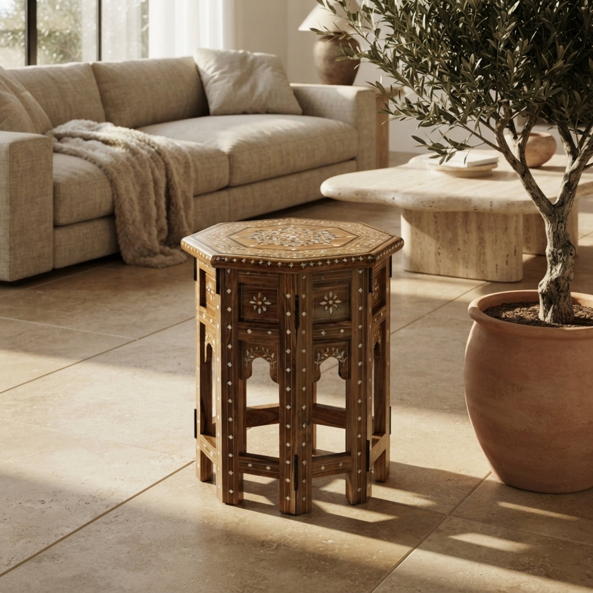A small octagonal wooden side table with carved floral details and brass hardware, placed in a sunlit living room beside a beige sofa and a potted olive tree.