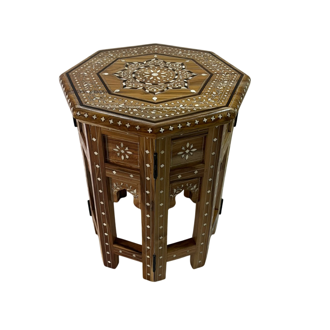 A small octagonal wooden side table with carved details stands beside a bed in a softly lit bedroom. A cream ceramic vase with white orchids rests on top.