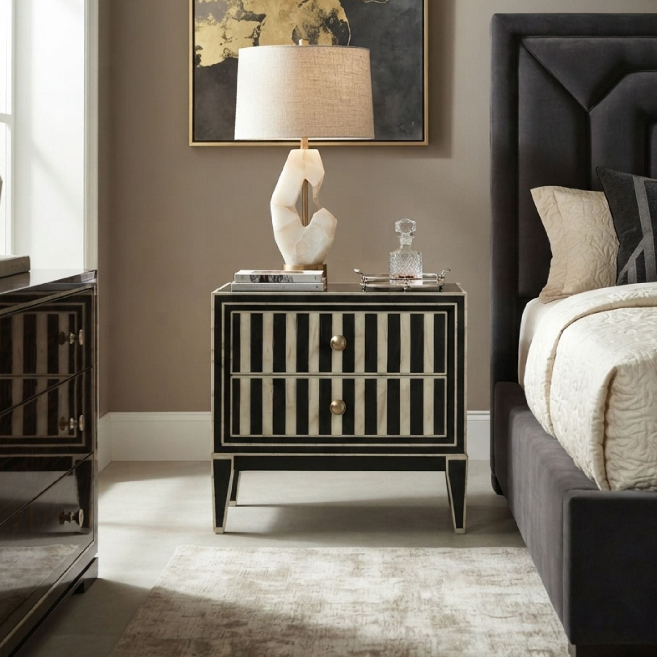 A black and beige striped nightstand in a styled bedroom setting, styled with a sculptural white lamp, books, and a glass decanter.