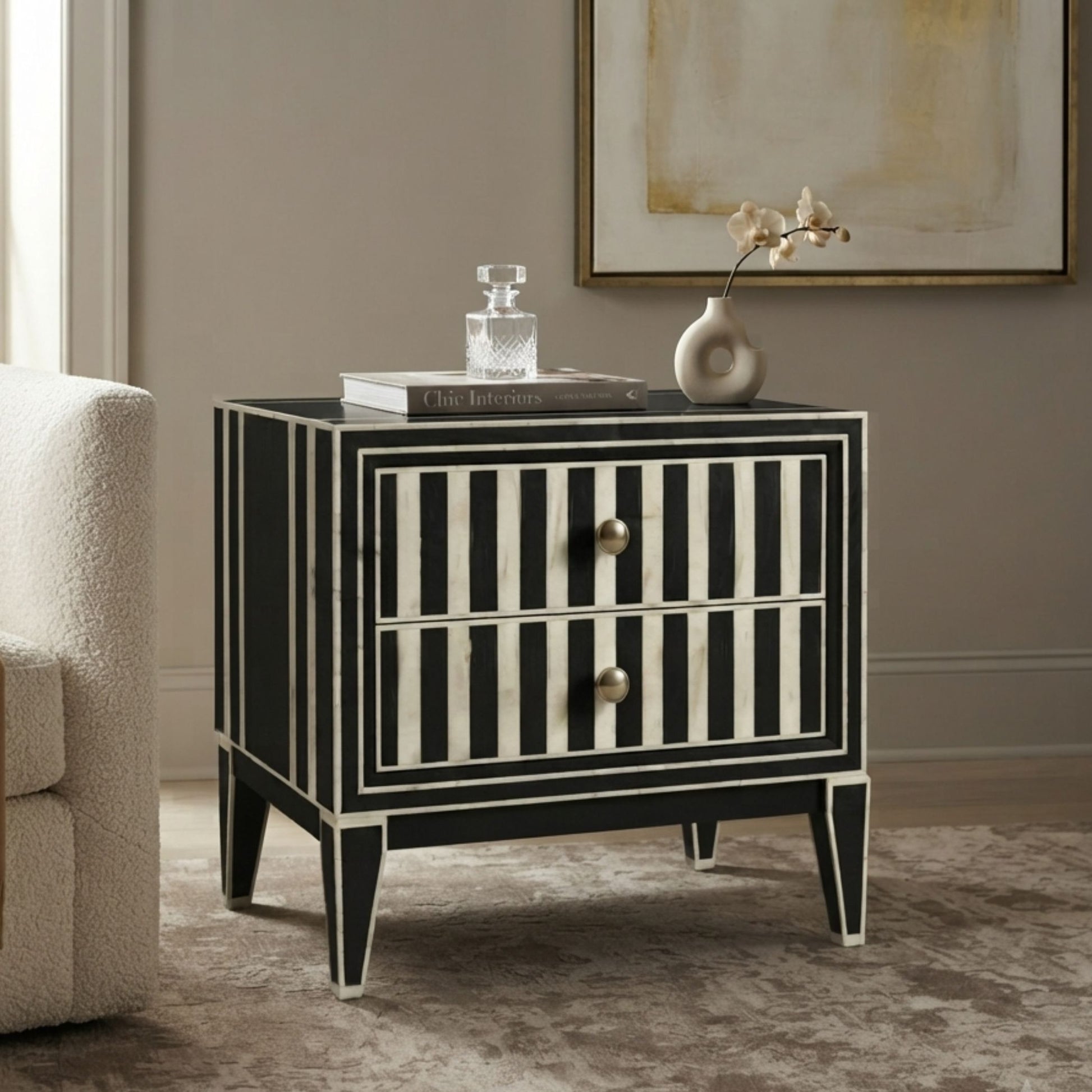 A black and white striped two-drawer nightstand with gold-tone knobs, styled in a living room setting next to a white armchair with a vase and books on top.