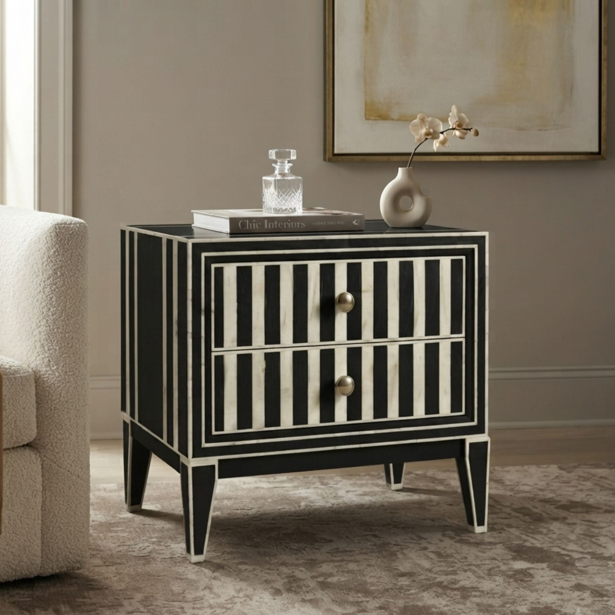 A black and white striped two-drawer nightstand with gold-tone knobs, styled in a living room setting next to a white armchair with a vase and books on top.