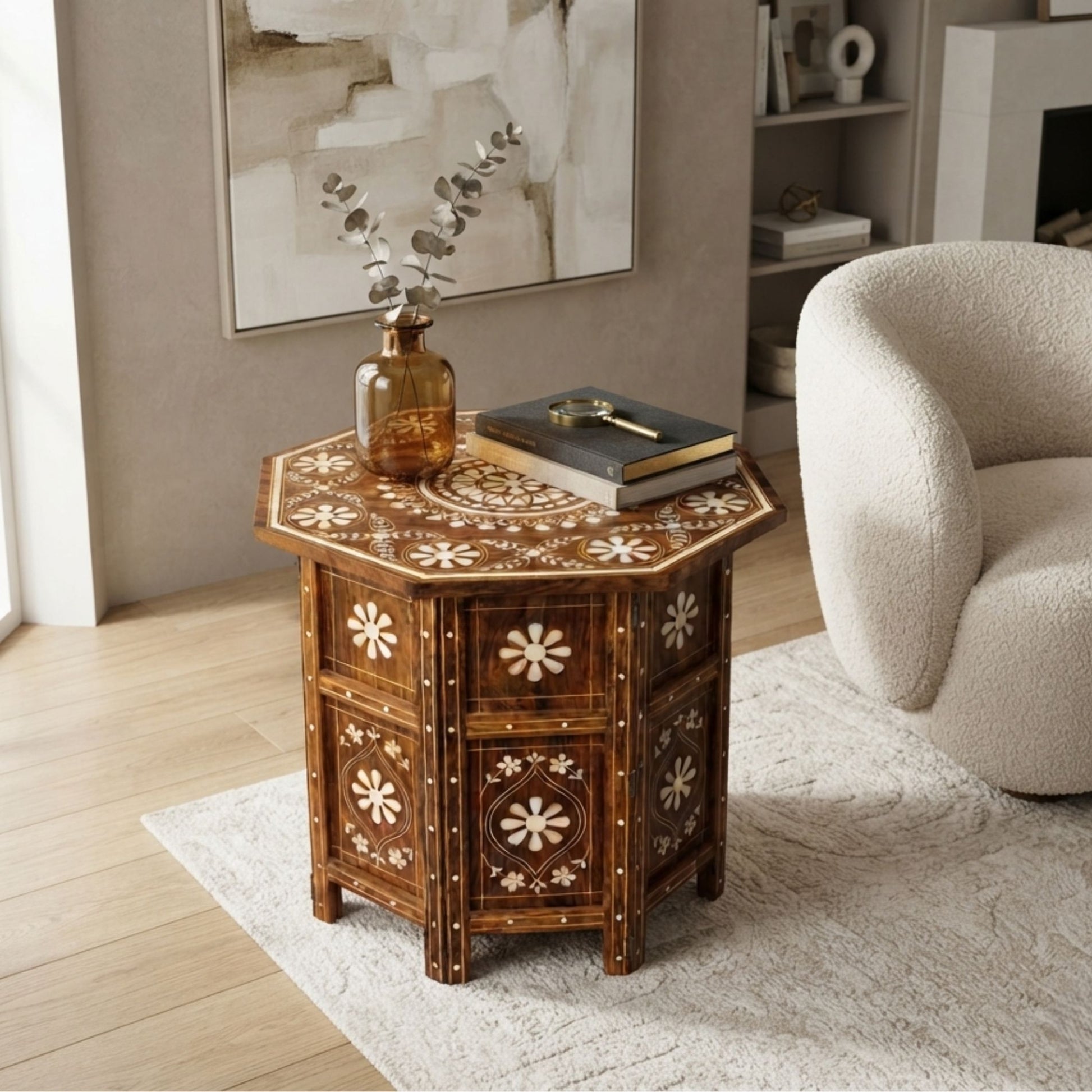 A hexagonal wooden side table with intricate floral inlay patterns, styled in a living room next to a cream-colored armchair with a glass vase and books on top.