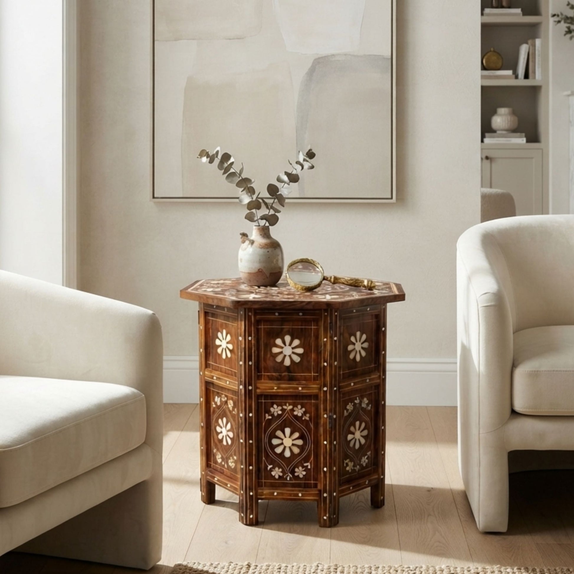 A dark brown octagonal wooden side table with intricate floral inlay details, styled in a bright living room setting between two cream-colored armchairs.