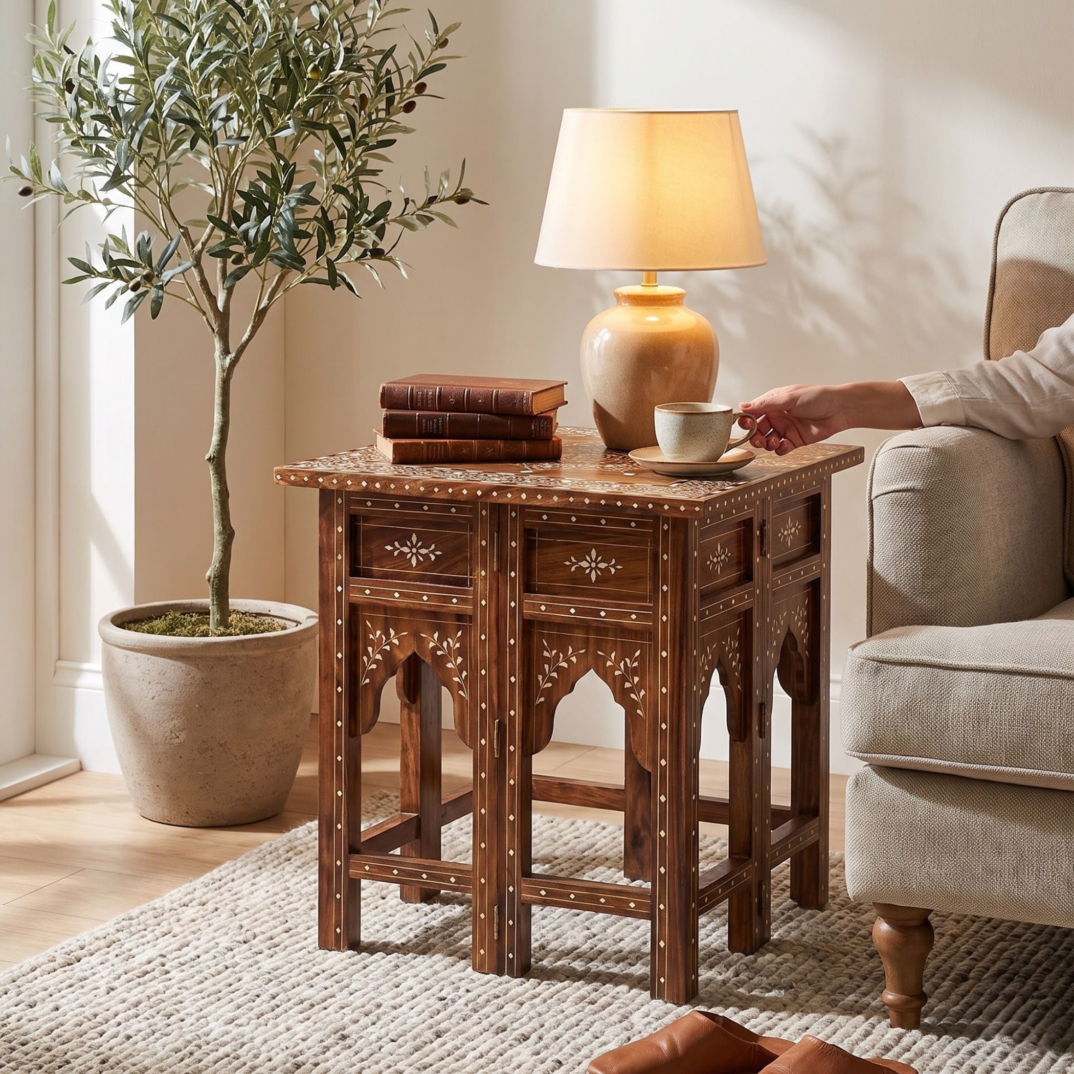 A dark brown carved wooden end table with intricate brass hardware and inlay details, styled in a cozy living room next to an armchair with a lamp and books on top.