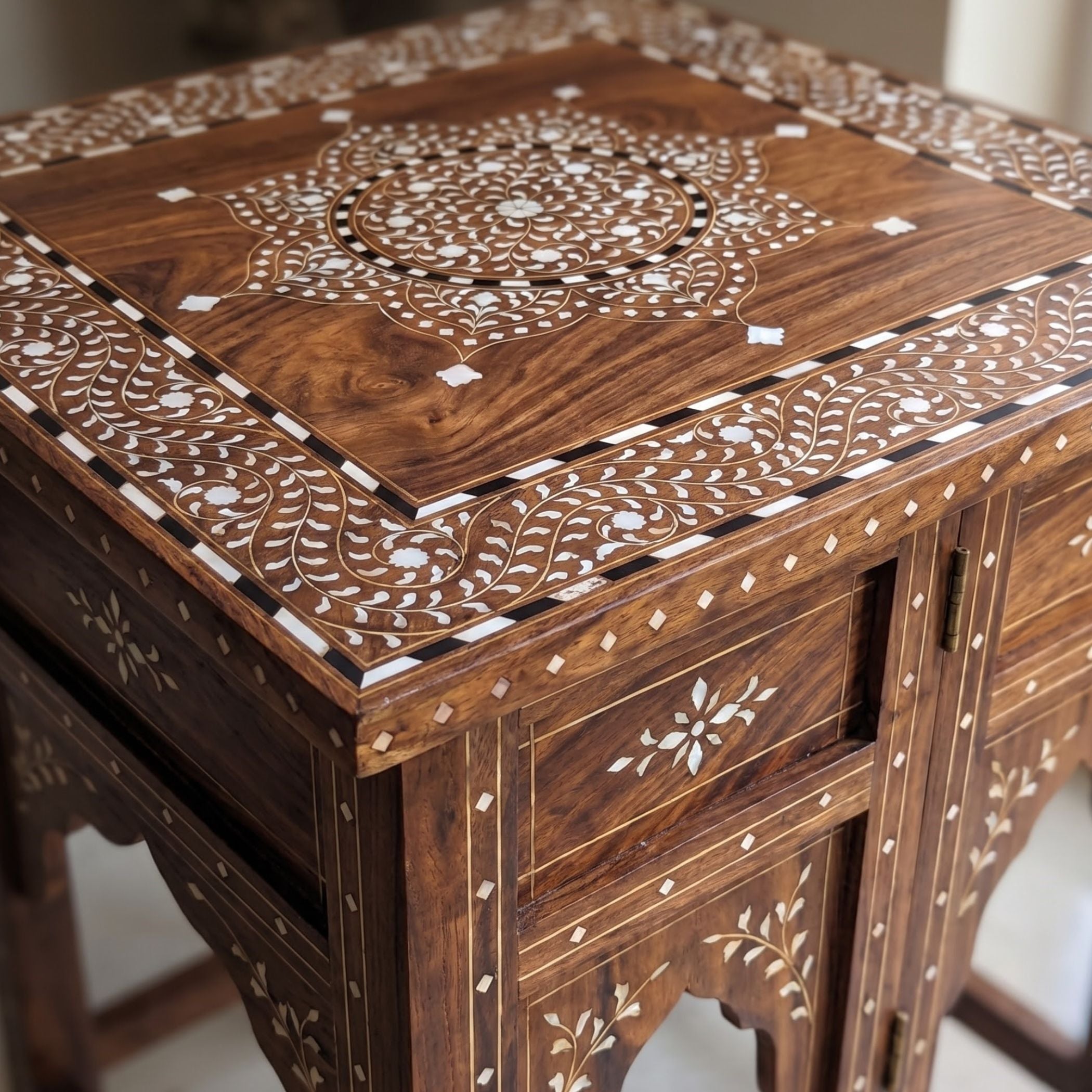 An ornate dark brown wooden side table decorated with intricate white and black bone inlay patterns on the tabletop and drawer fronts.