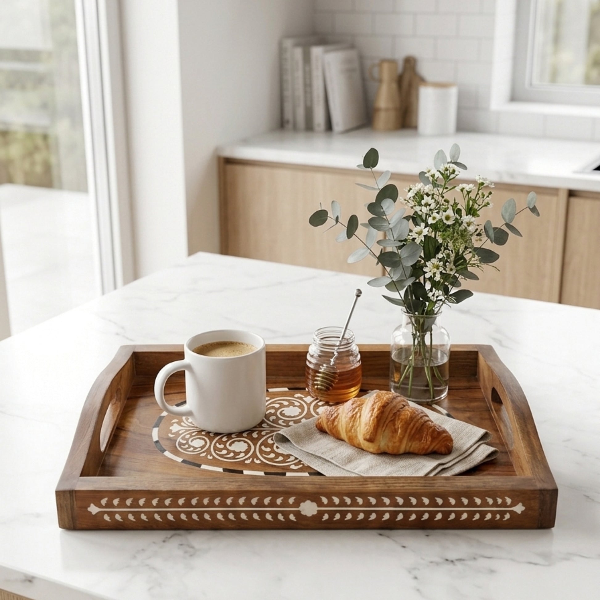 A rectangular wooden serving tray with carved geometric details and side handles, styled on a white marble kitchen counter with a white mug, honey jar, croissant, and a small vase of greenery.