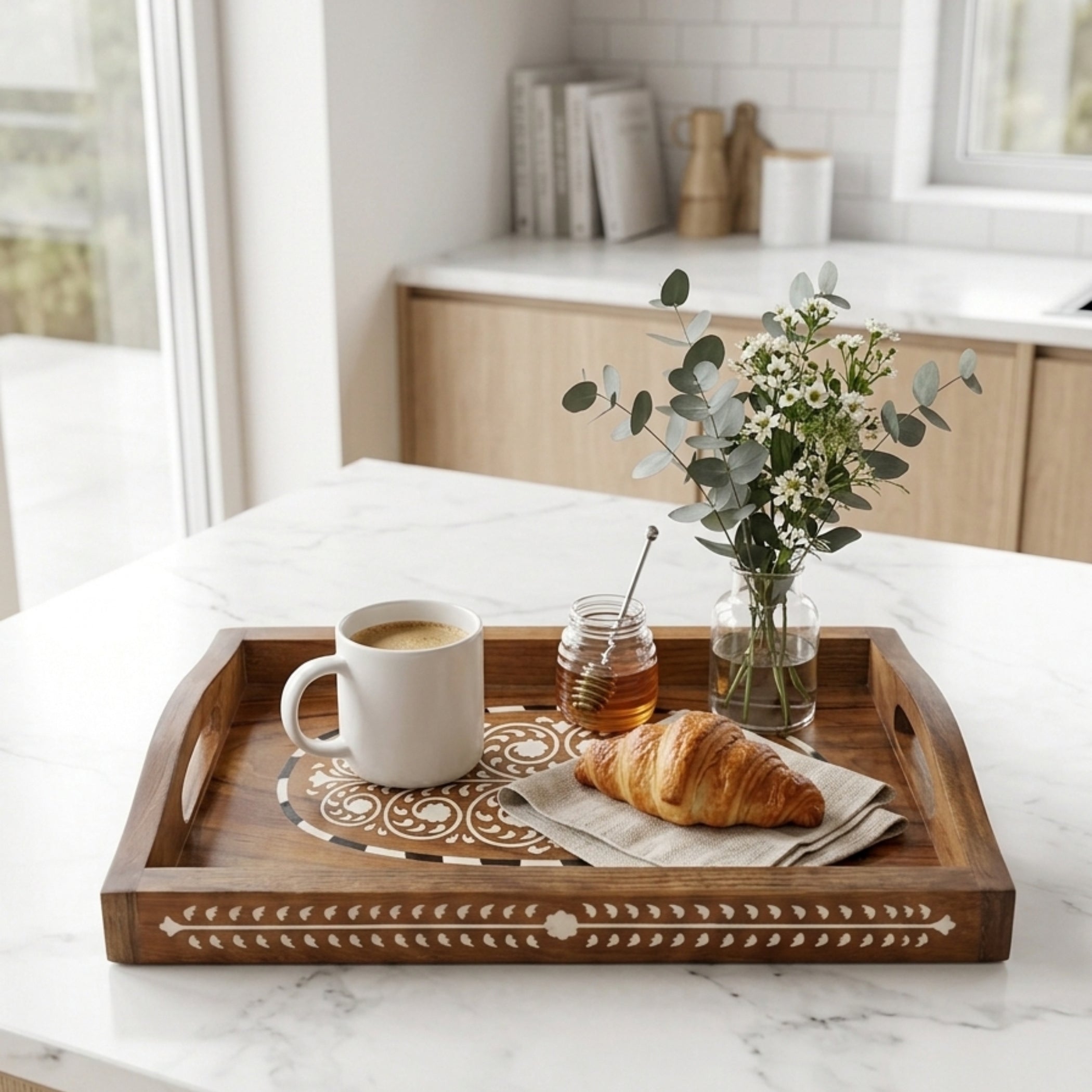 A rectangular wooden serving tray with carved geometric details and side handles, styled on a white marble kitchen counter with a white mug, honey jar, croissant, and a small vase of greenery.